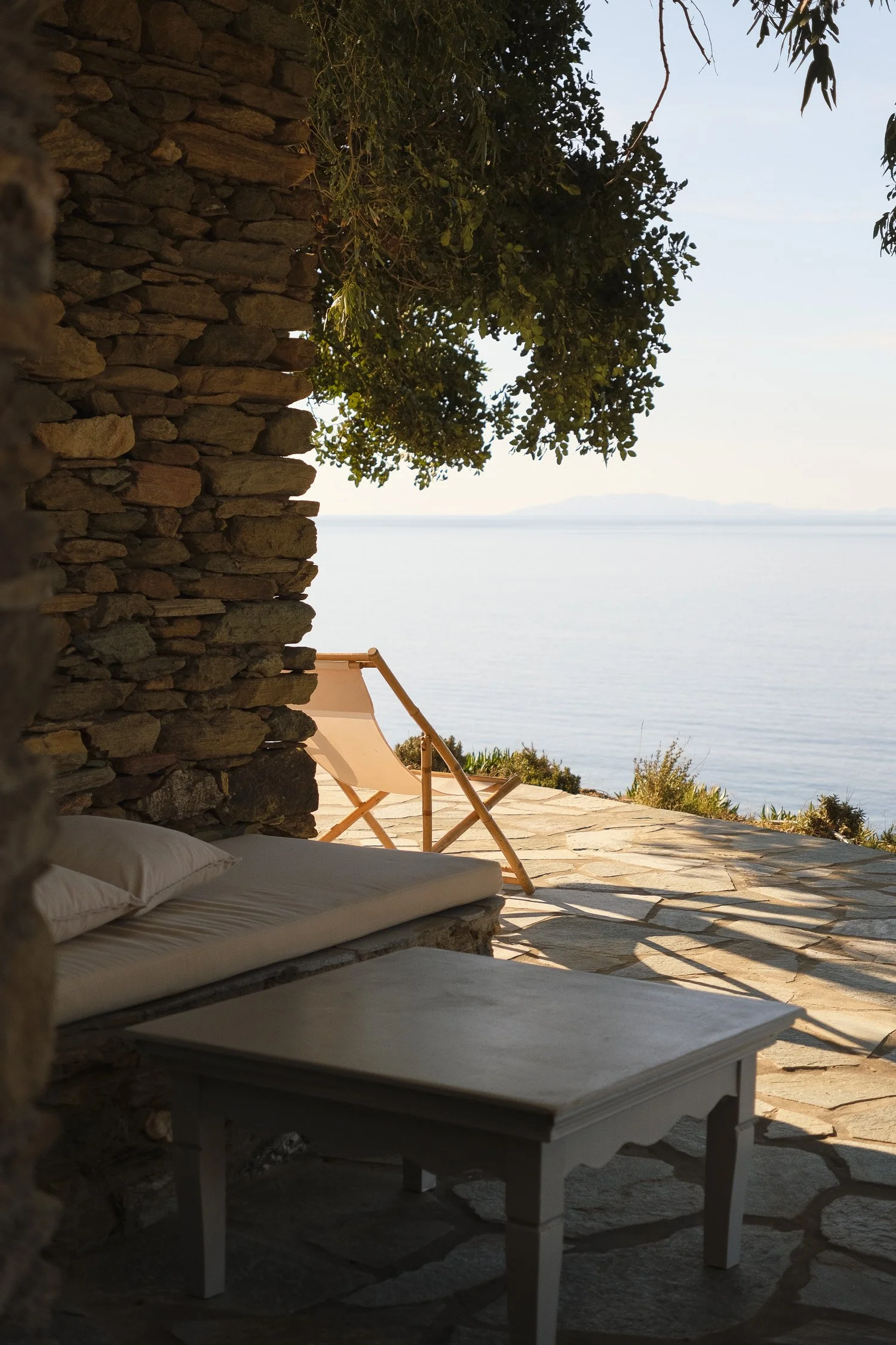 A cozy outdoor seating area with a stone wall, white cushion, and a small table, overlooking the water with a lounge chair and tropical trees.