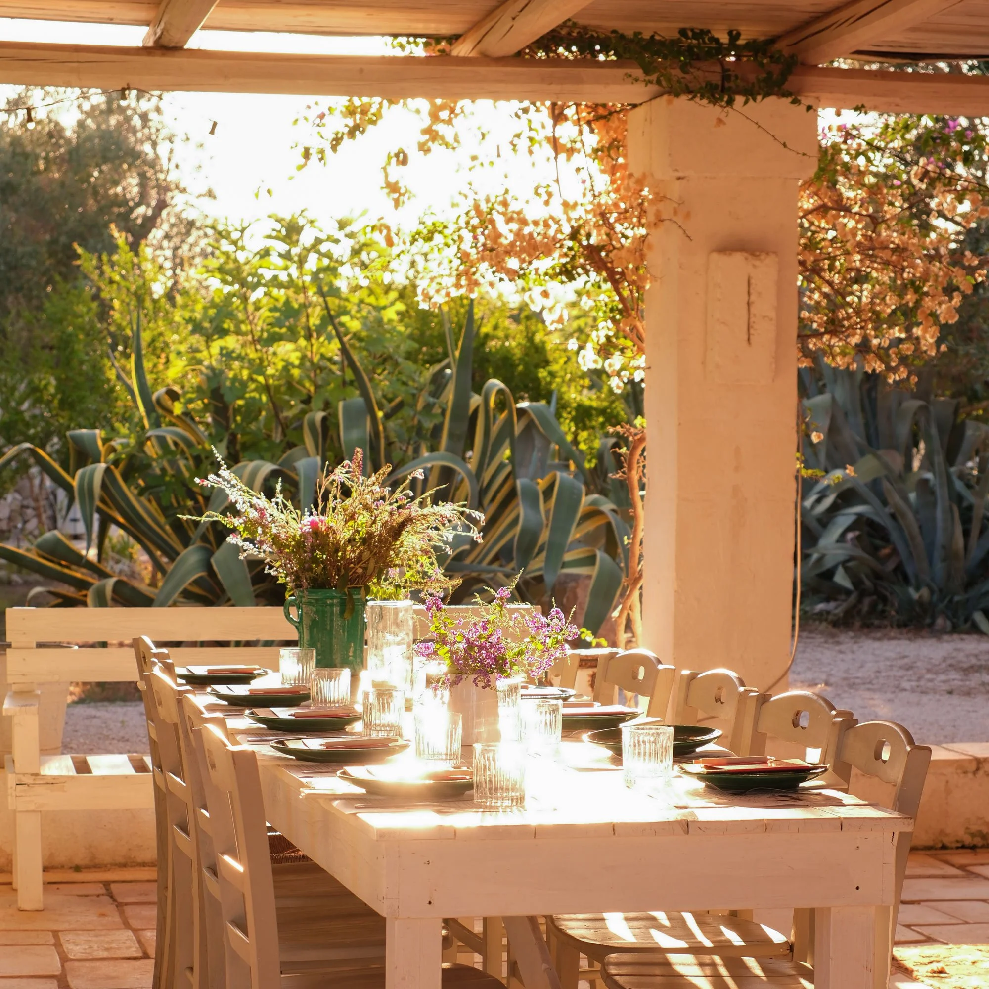 Outdoor dining table with place settings and floral centerpieces, on a patio with greenery in the background
