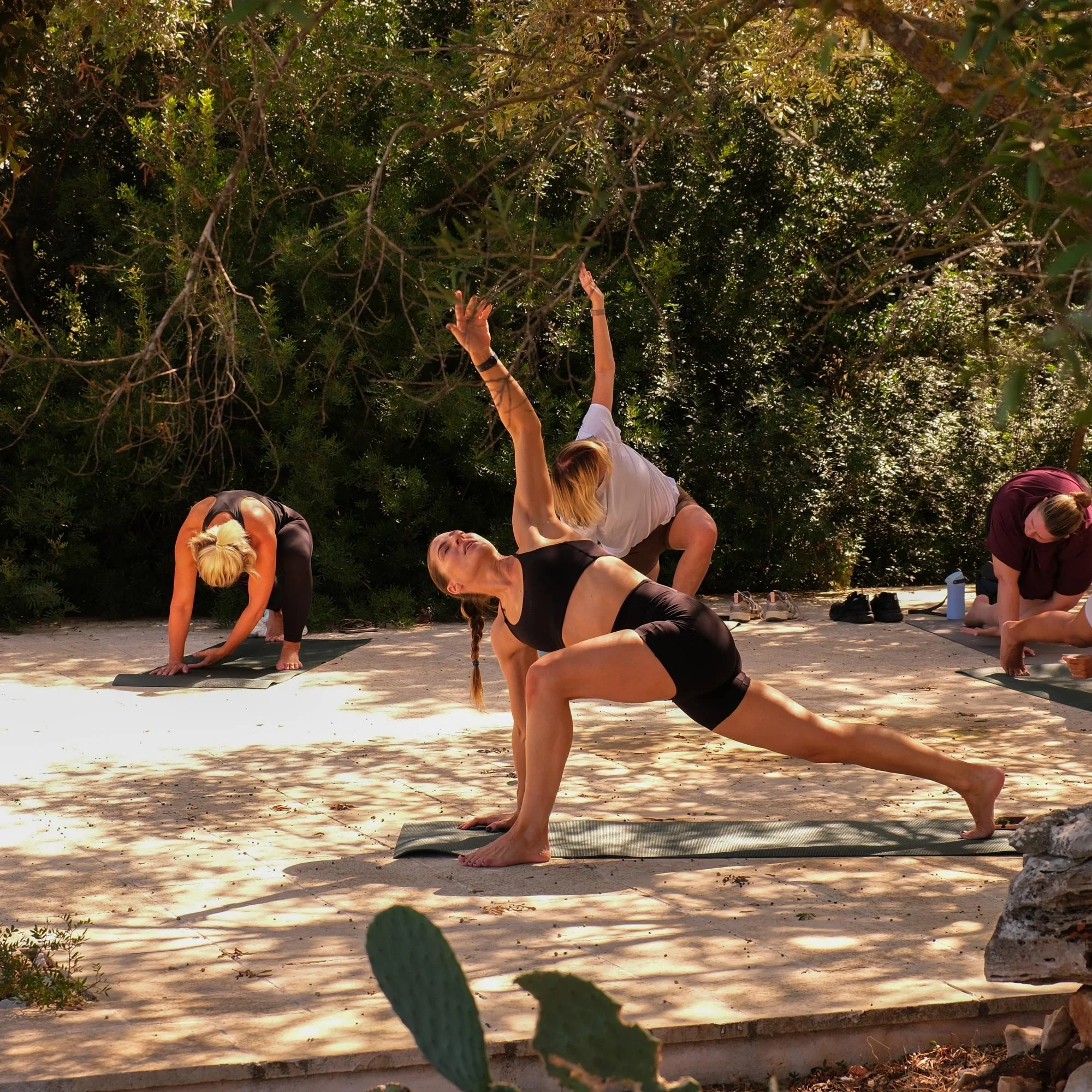 Women practicing outdoor yoga on mats in a shaded natural setting with trees and bushes.