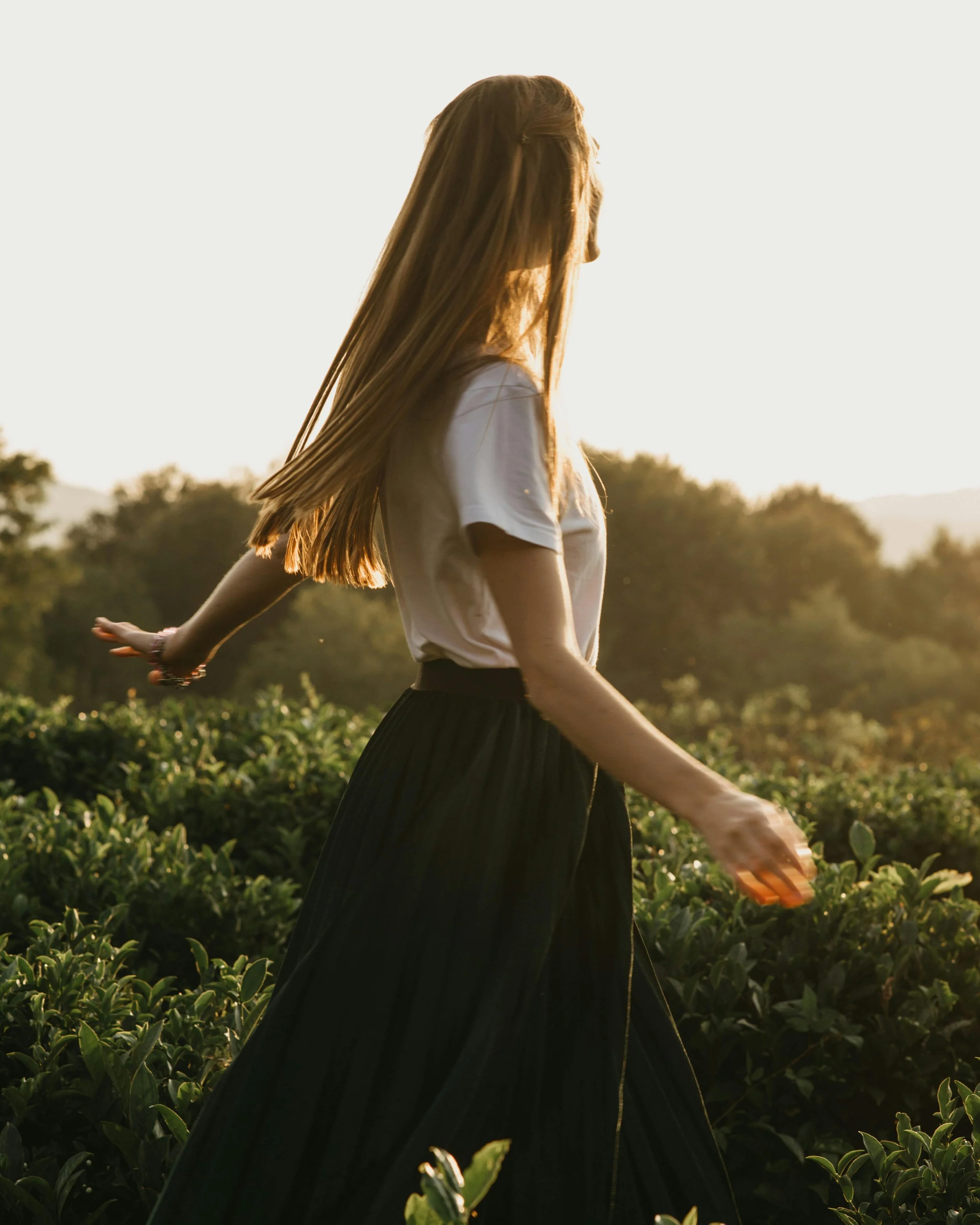 A woman with long hair walking through a lush green field at sunset.