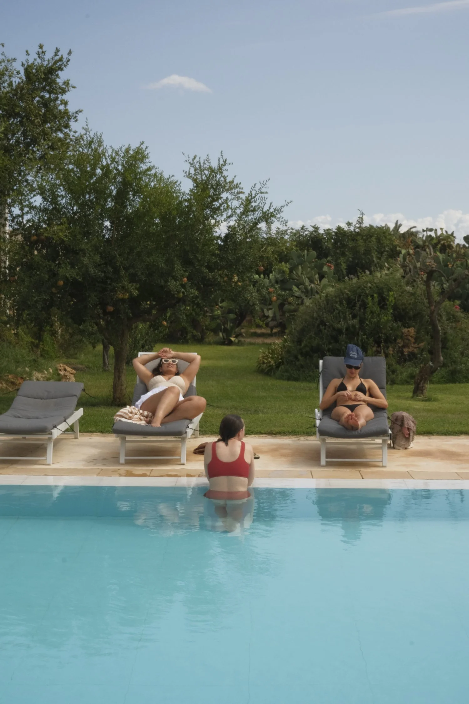 People relaxing by a swimming pool on lounge chairs outdoors with trees and blue sky in the background.