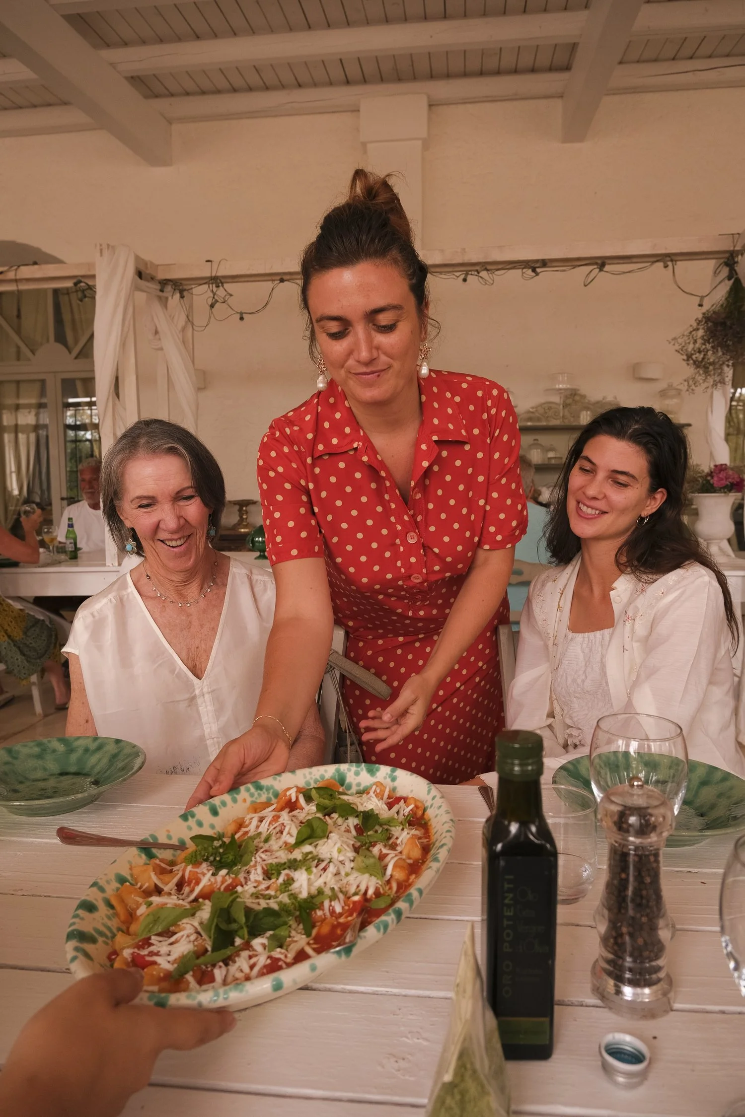 Three women sitting at a dining table with Italian dish of pasta with tomato sauce, cheese, and basil; a bottle of olive oil, pepper grinder, and wine glass are on the table, with one woman serving the dish.