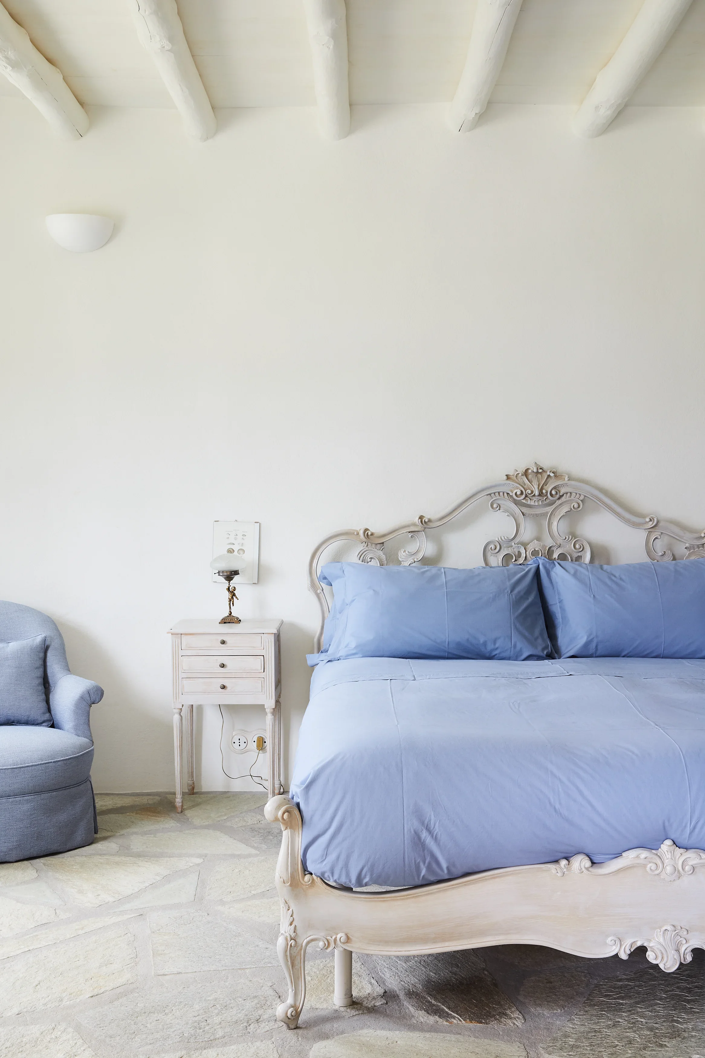 A bedroom with a vintage white bed frame, blue bedding, a small white nightstand, a grey armchair with a cushion, and a white wall with a wall-mounted light fixture.