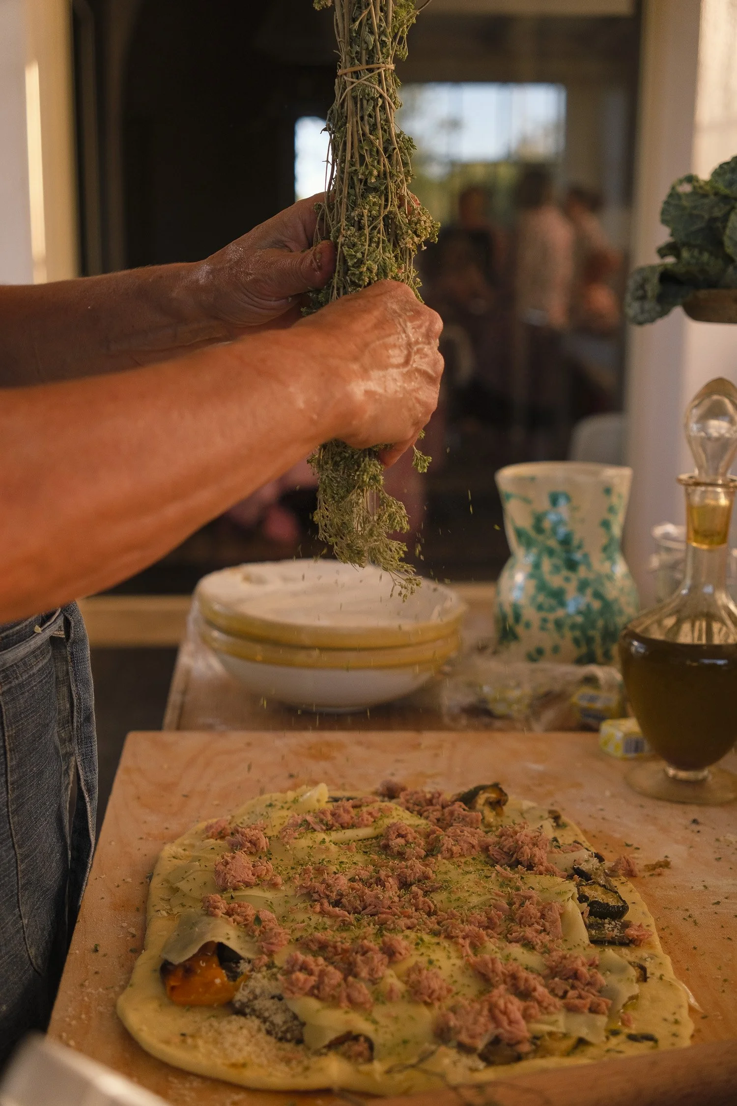Person seasoning a homemade pizza with herbs and ground meat, with people in the background in a kitchen or dining area.