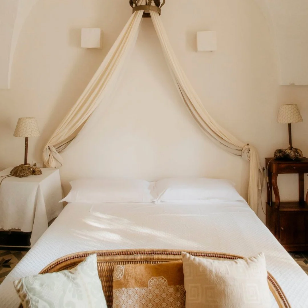 A cozy bedroom with an off-white bed adorned with decorative pillows and a draped canopy above the headboard. Wooden nightstands with lamps flank the bed, and there are white wall-mounted light fixtures behind the canopy.