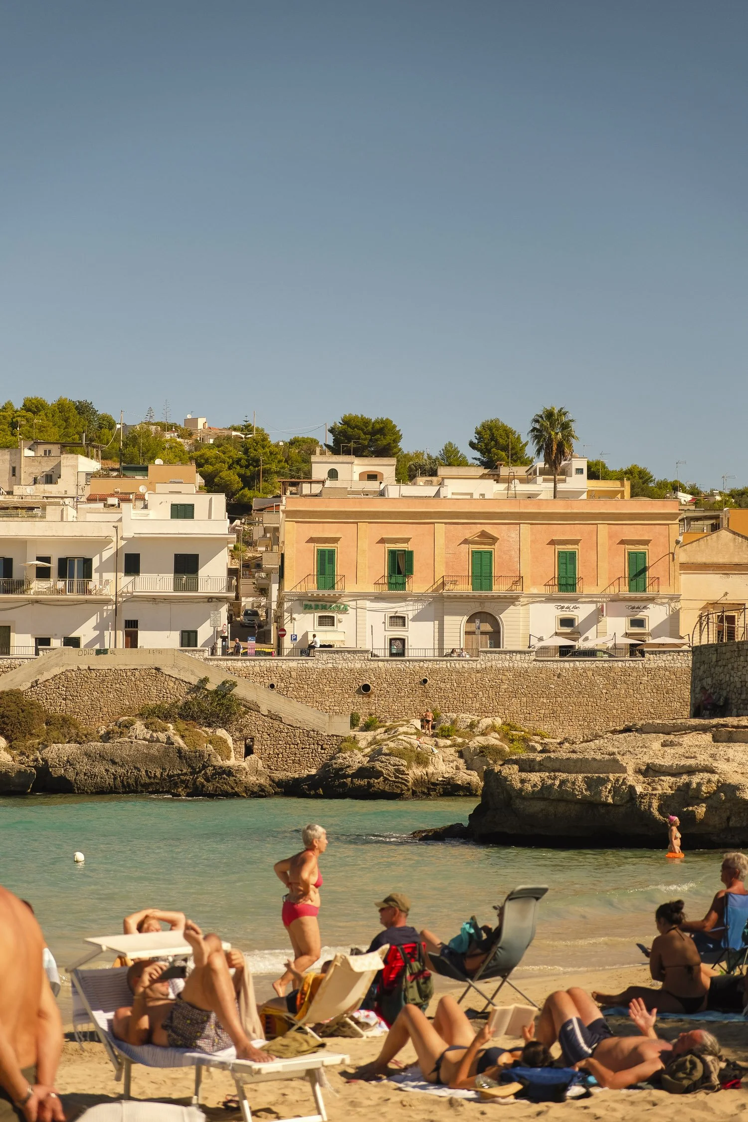 Beach scene with people lounging on chairs, a woman in a red swimsuit standing in the water, and colorful buildings with green shutters in the background under a clear blue sky.