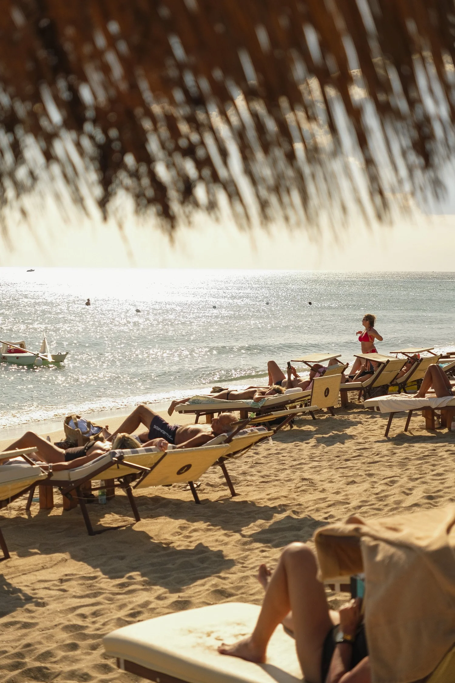 Beach scene with lounge chairs, people sunbathing, and a woman standing on the sand, with the ocean and boats in the background, viewed under a thatched roof.