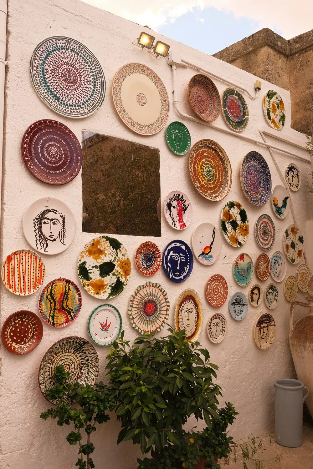 Decorative ceramic plates with colorful patterns and face illustrations on a white wall, with potted plant and large vase below.