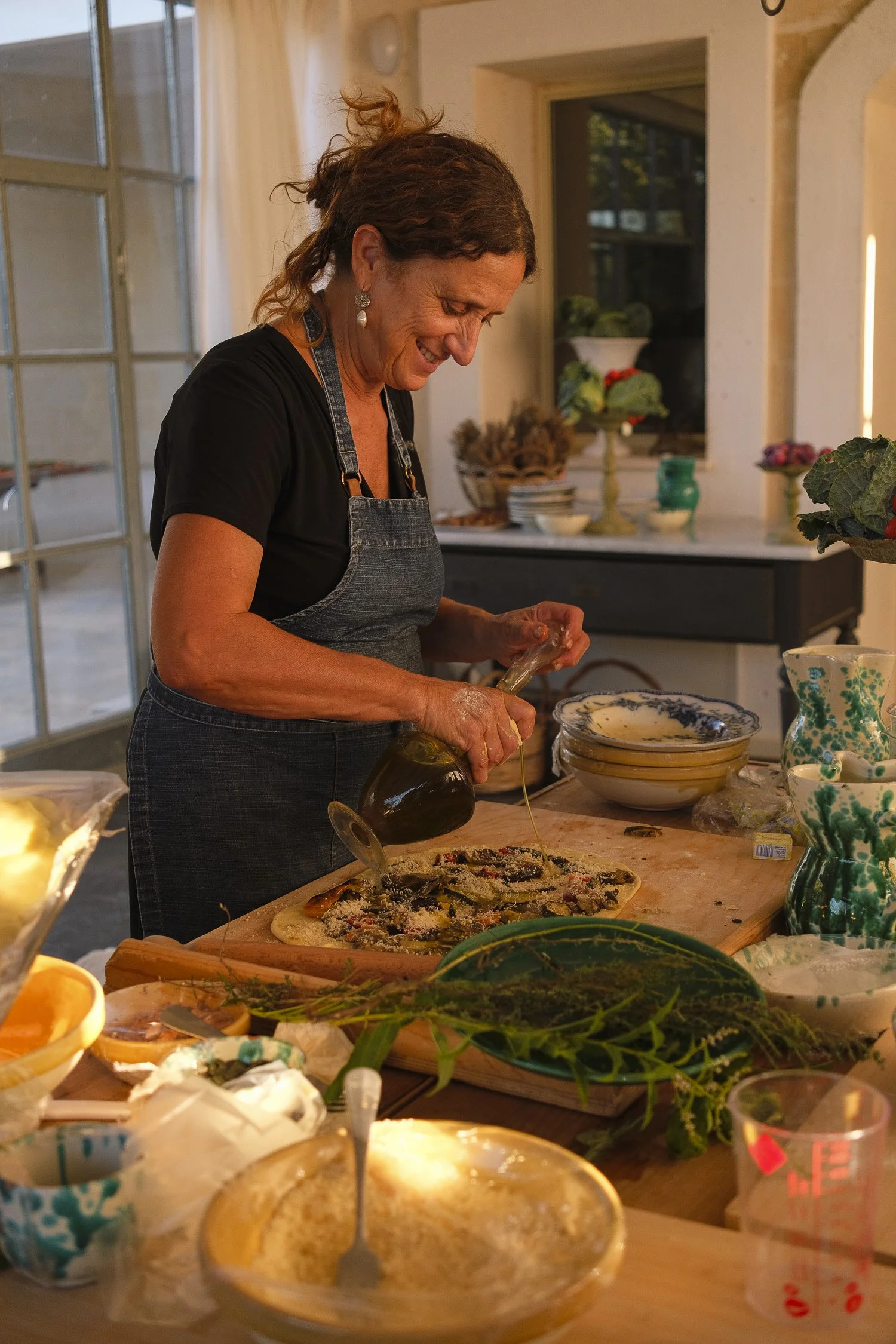 A woman preparing a pizza in a cozy kitchen, pouring olive oil onto it, surrounded by kitchen utensils, dishes, and greenery.