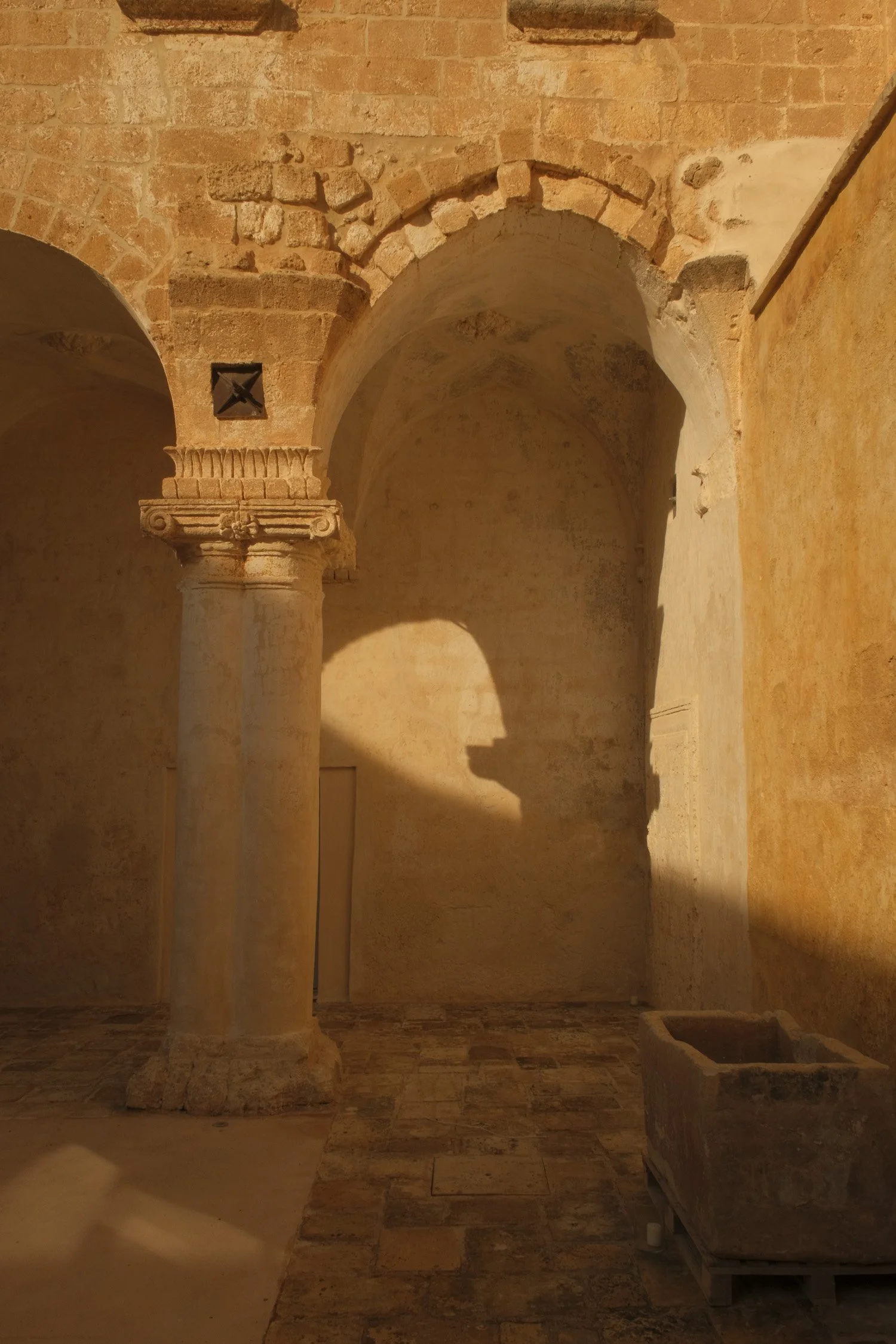 Ancient stone interior with a column, archway, and shadowed wall, with a stone basin on a wooden stand.