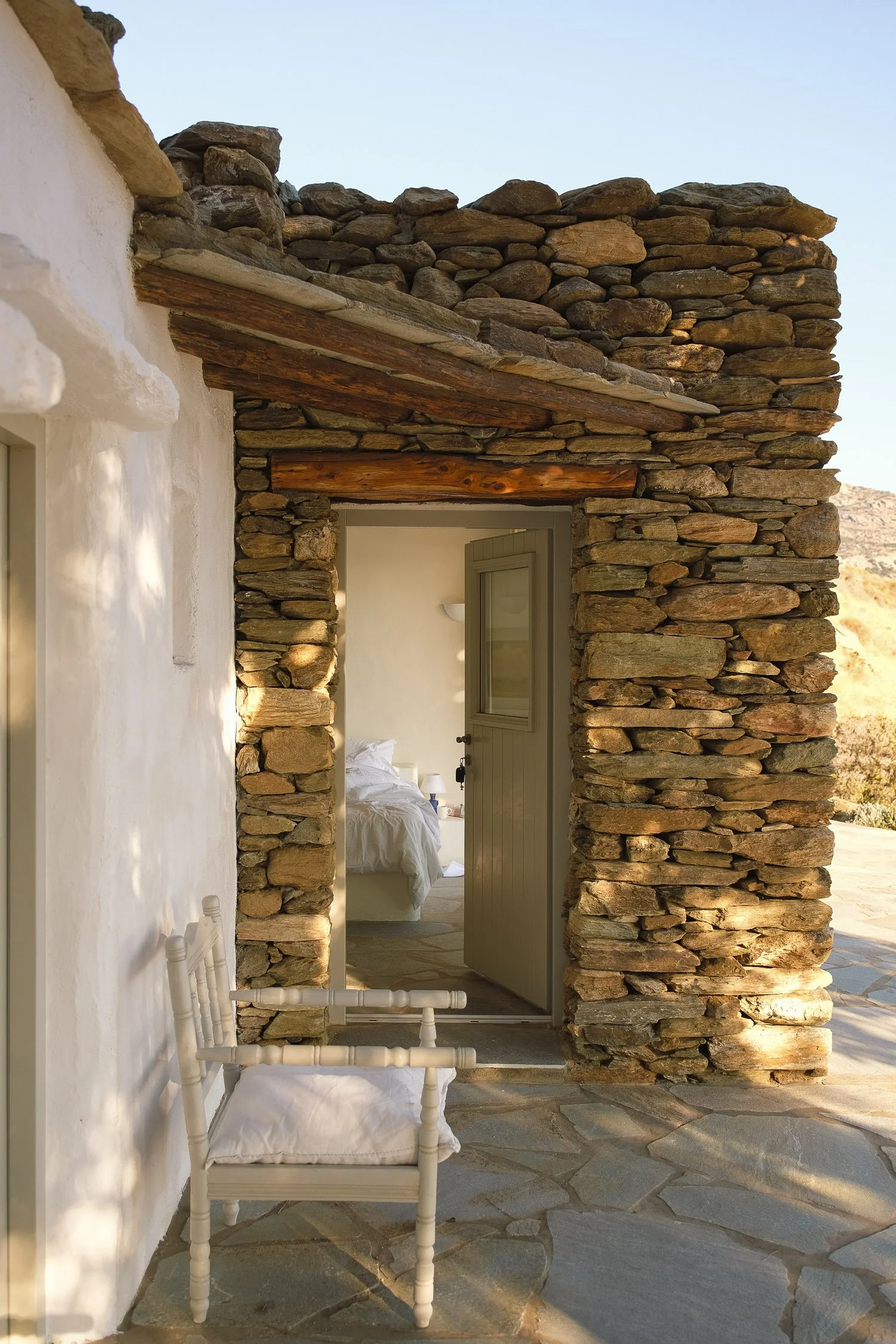 A view of a rustic stone and wood exterior wall leading into a bedroom with a partially open door, white bed, and bedside table, outdoor patio with a white chair.