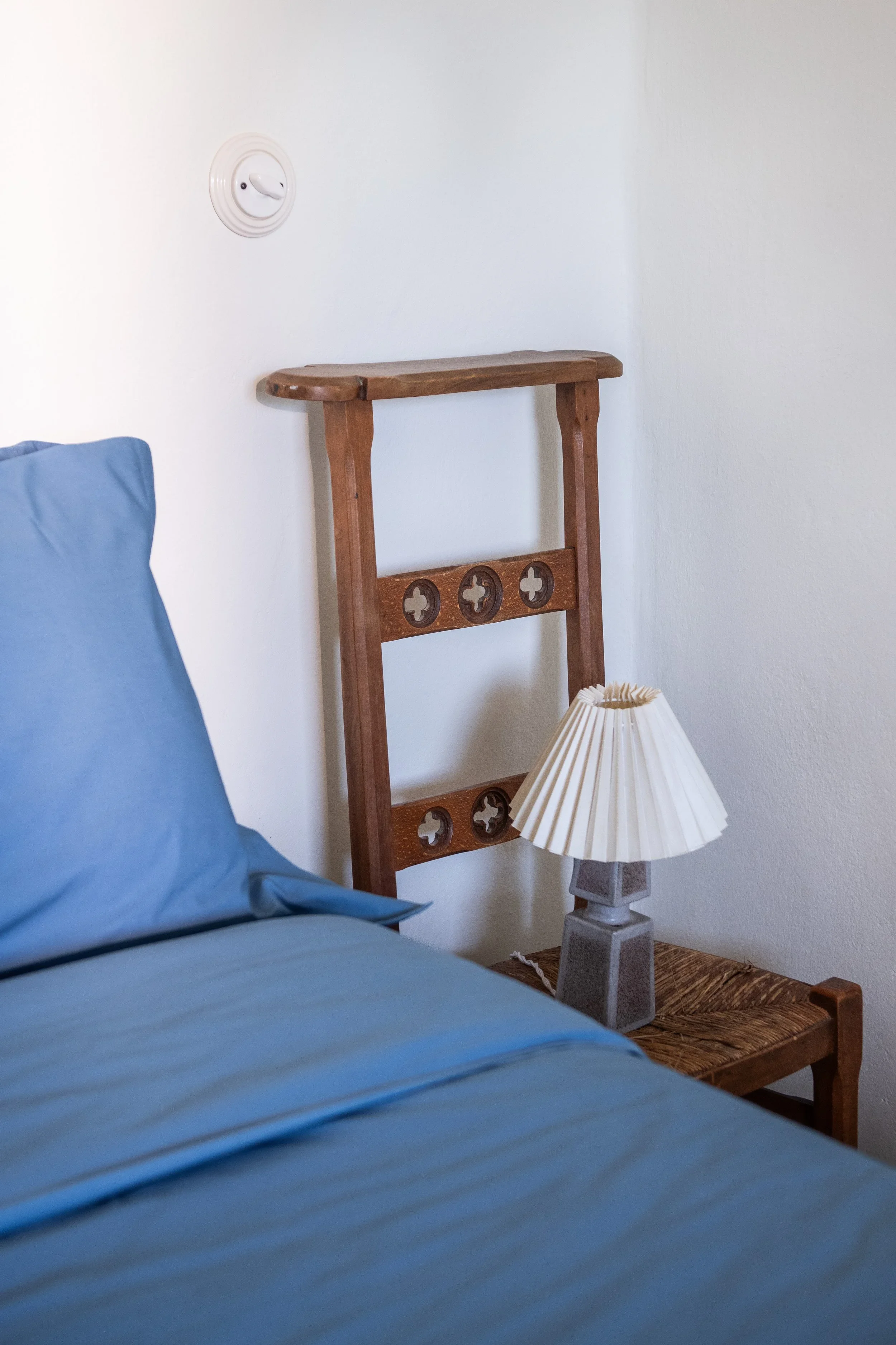 A bedroom corner with a bed in blue sheets, a wooden nightstand with a table lamp, and a wooden wall decor piece above the nightstand.