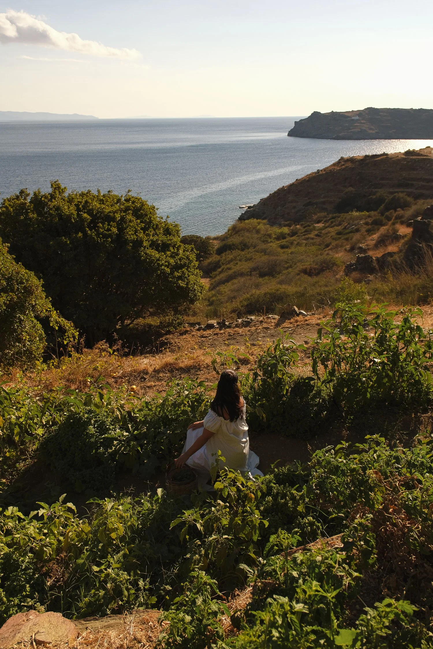 A woman in a white dress sits in a lush, green garden overlooking a coastal landscape with hills, trees, and an ocean under a partly cloudy sky.
