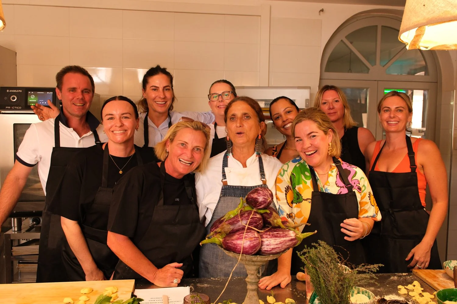A group of smiling people in a kitchen, wearing aprons, gathered around a chef holding a bowl of eggplants.