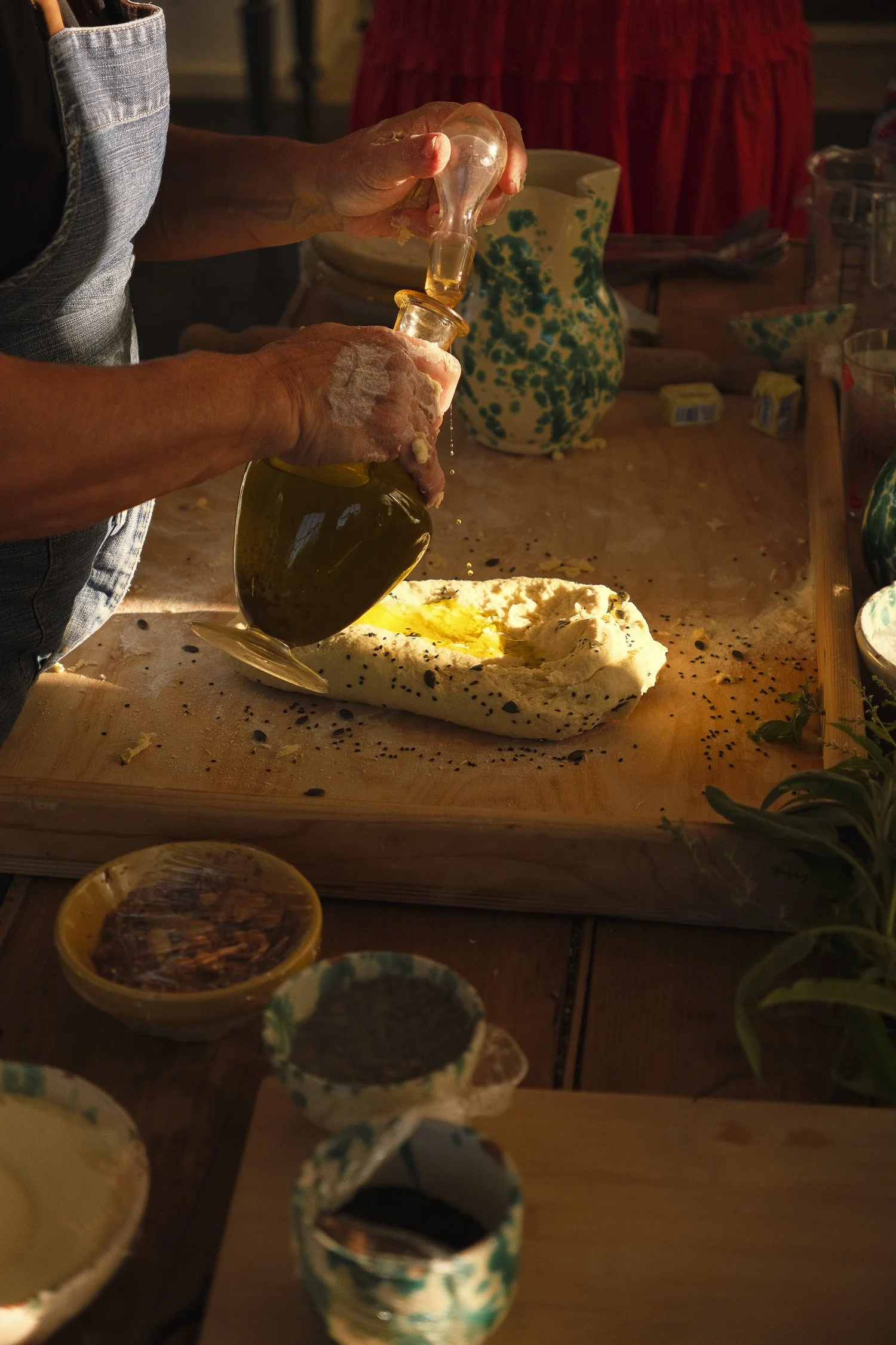 Person preparing bread with olive oil on a cutting board, various bowls with condiments, pottery, and herbs. Sunlight illuminating the scene.