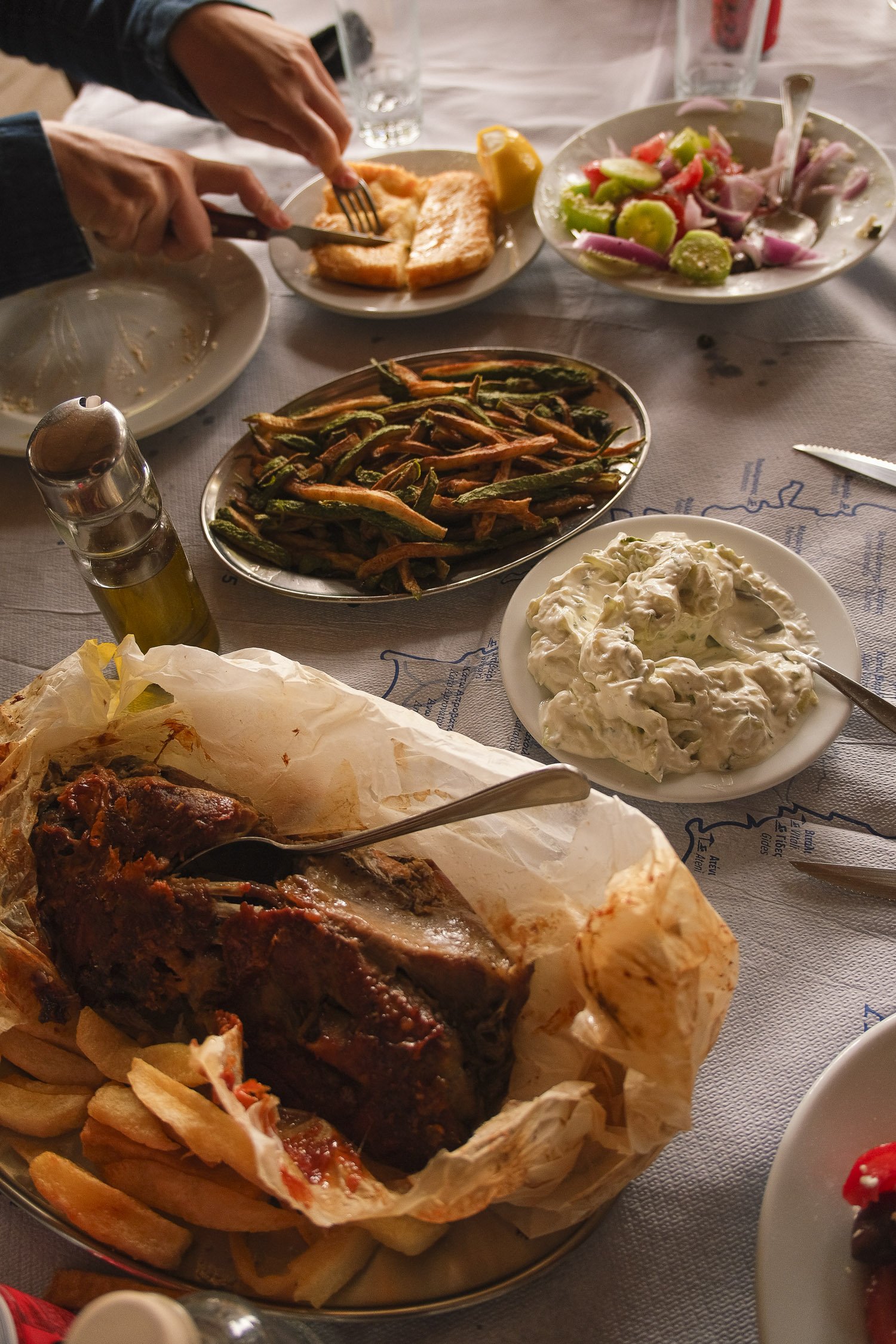 A table filled with various dishes including a roasted meat served on parchment paper with fries, a bowl of potato salad, a plate of green beans and carrots, a bowl of colorful vegetable salad, a plate with breaded fish fillet, and a container of olive oil and vinegar. Hand holding a fork and knife cutting the fried fish.