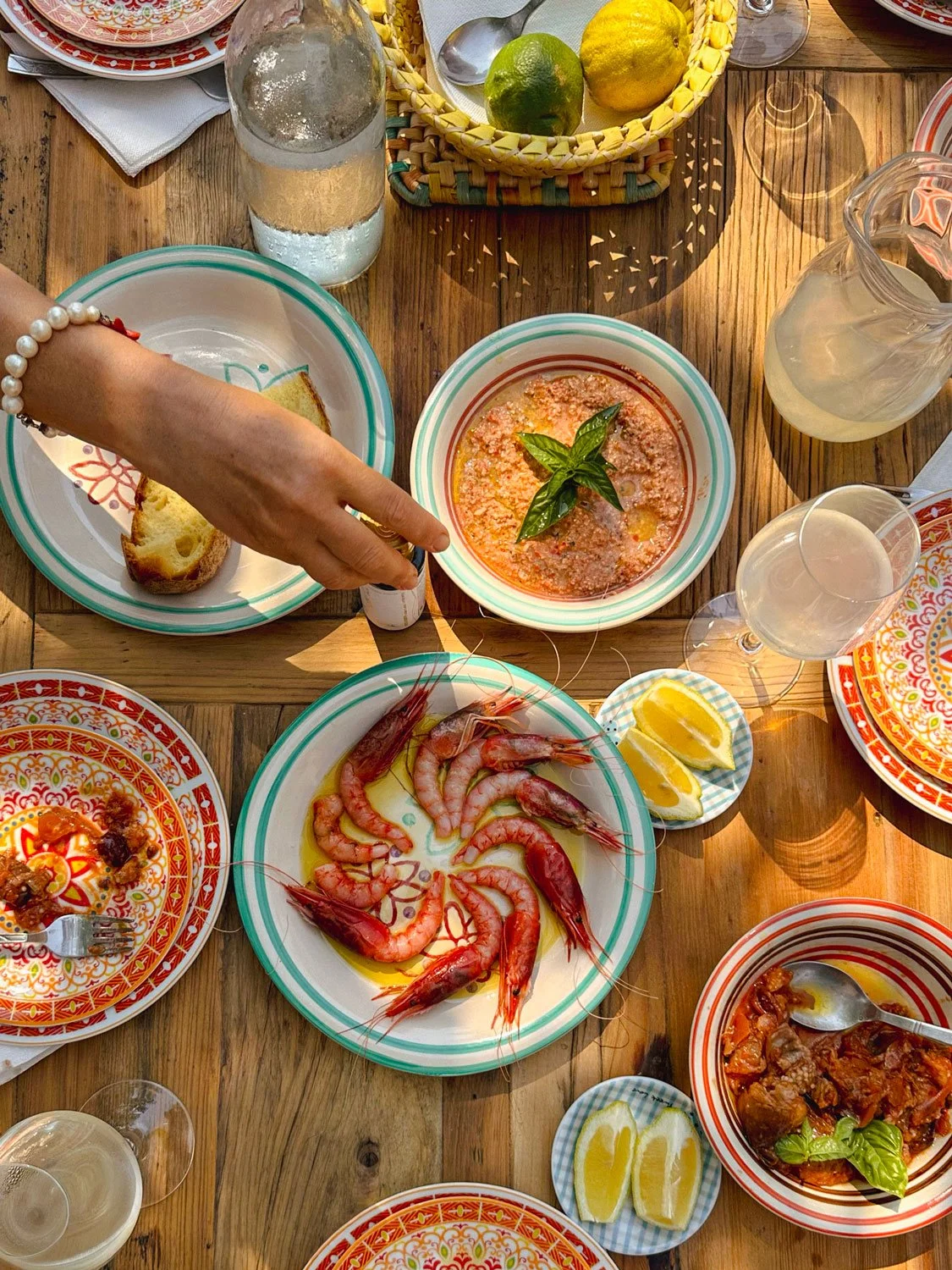A top-down view of a seafood feast on a wooden table, featuring shrimp, lemon wedges, a bowl of pink sauce with basil, a plate of bread, a glass of water, and other dishes with colorful patterns.
