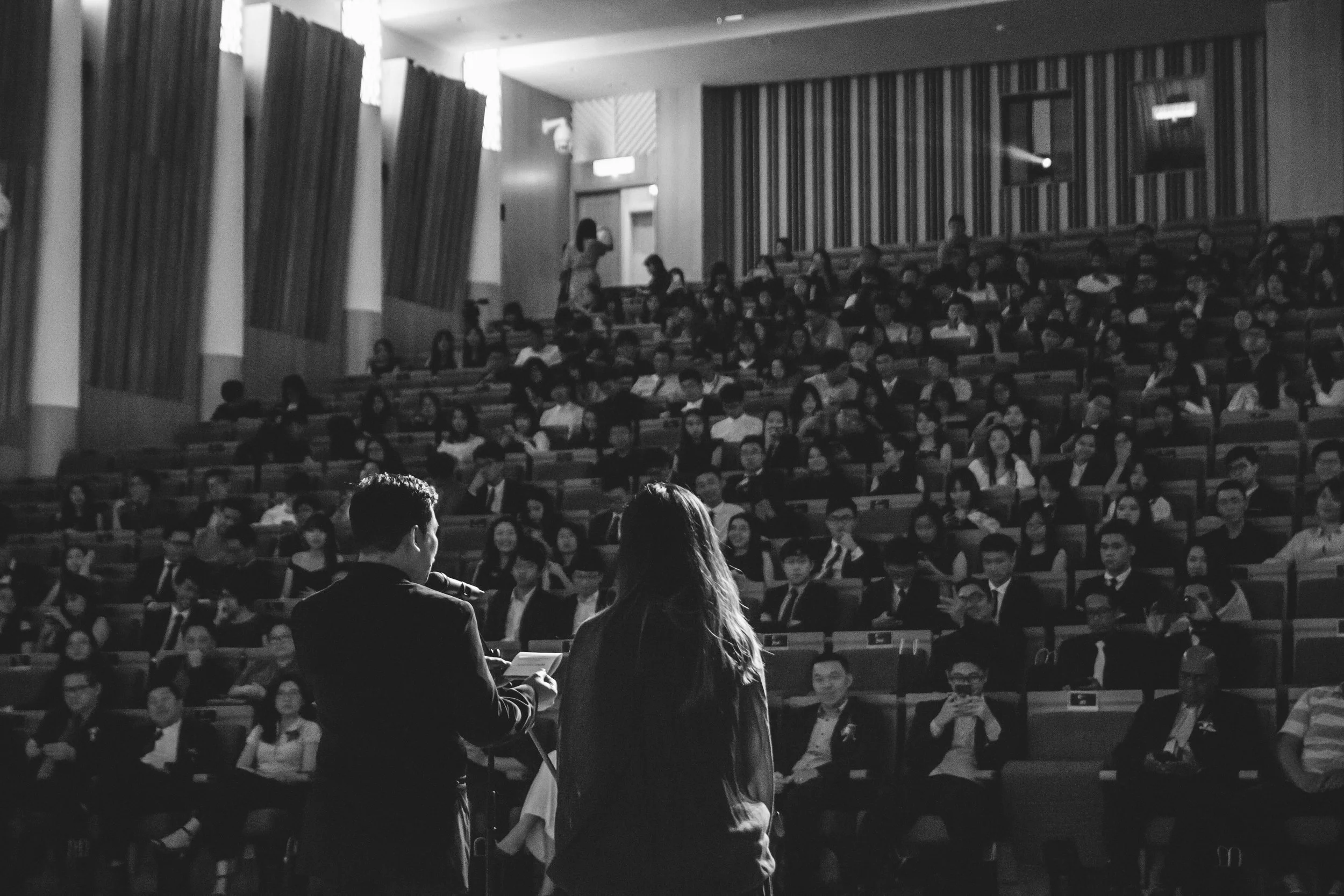 A black and white photo of a large auditorium filled with seated audience members. In the foreground, a man and a woman are standing in front of the audience, possibly speaking or presenting. The woman is holding a piece of paper. The audience members are attentively listening, with some taking notes or looking forward. There are two people on the stage near the speakers, and the auditorium has high windows with curtains and wooden wall panels.