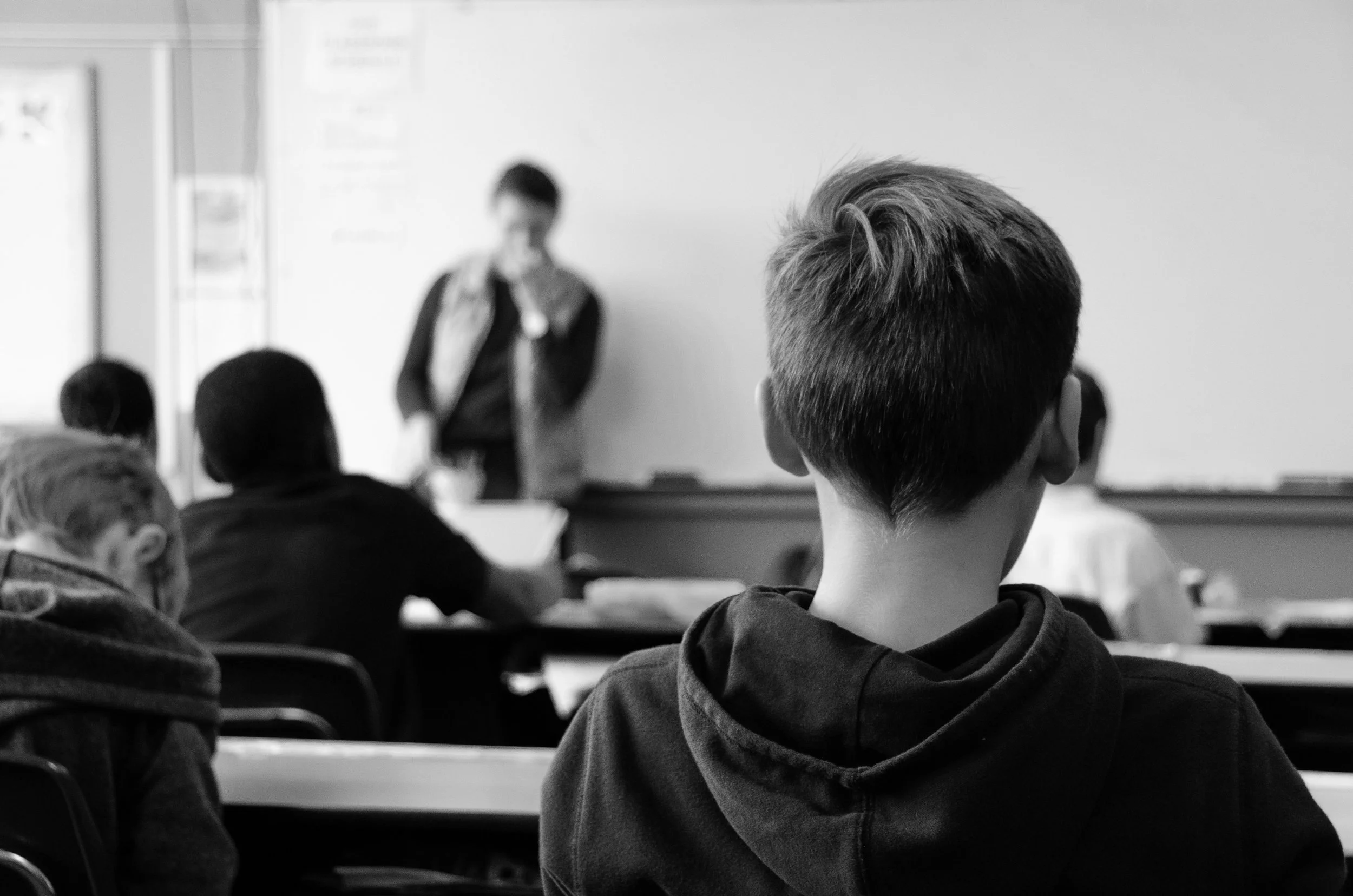 A classroom scene black and white photo showing students seated at desks facing a teacher standing at the front near a whiteboard, with a student in the foreground.