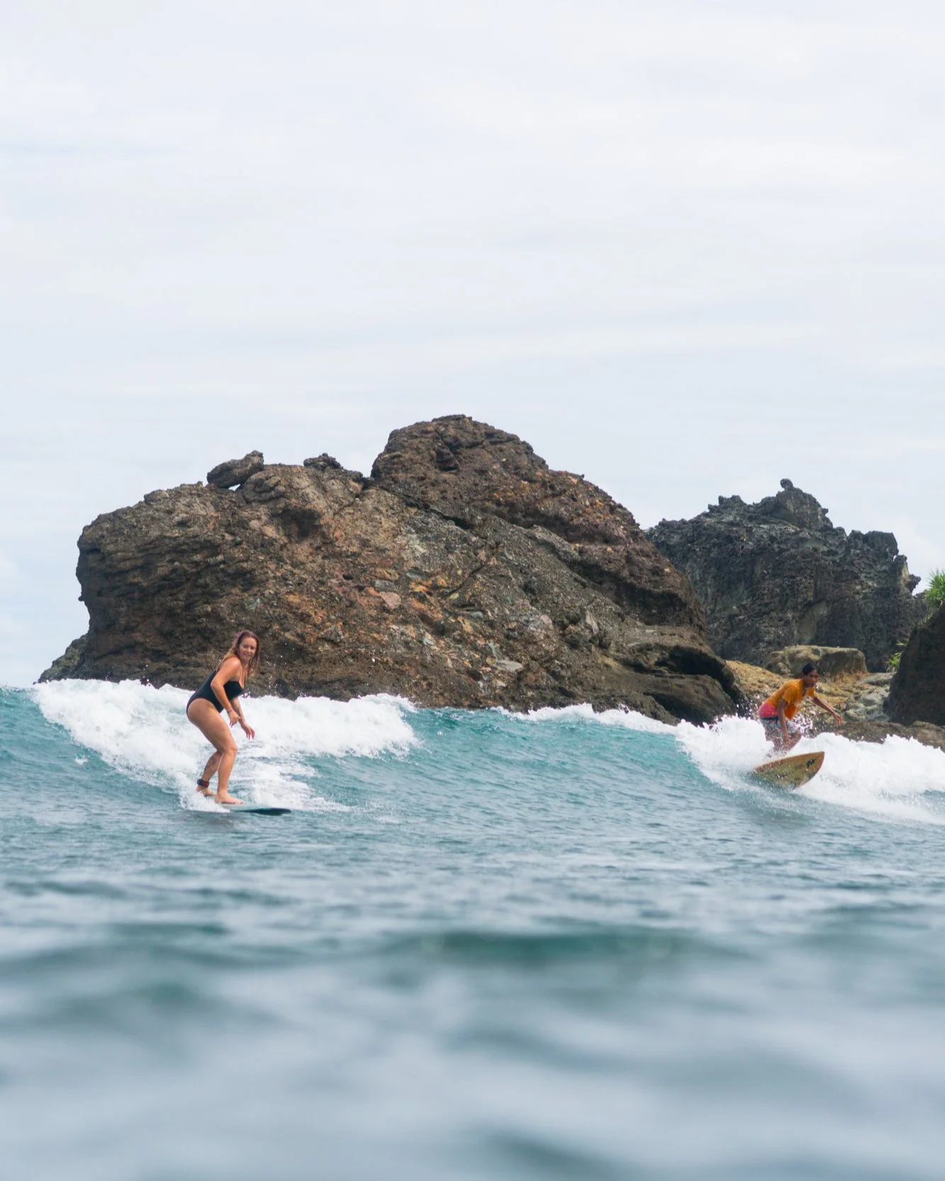 Two women surfing near rocky shoreline on a cloudy day.