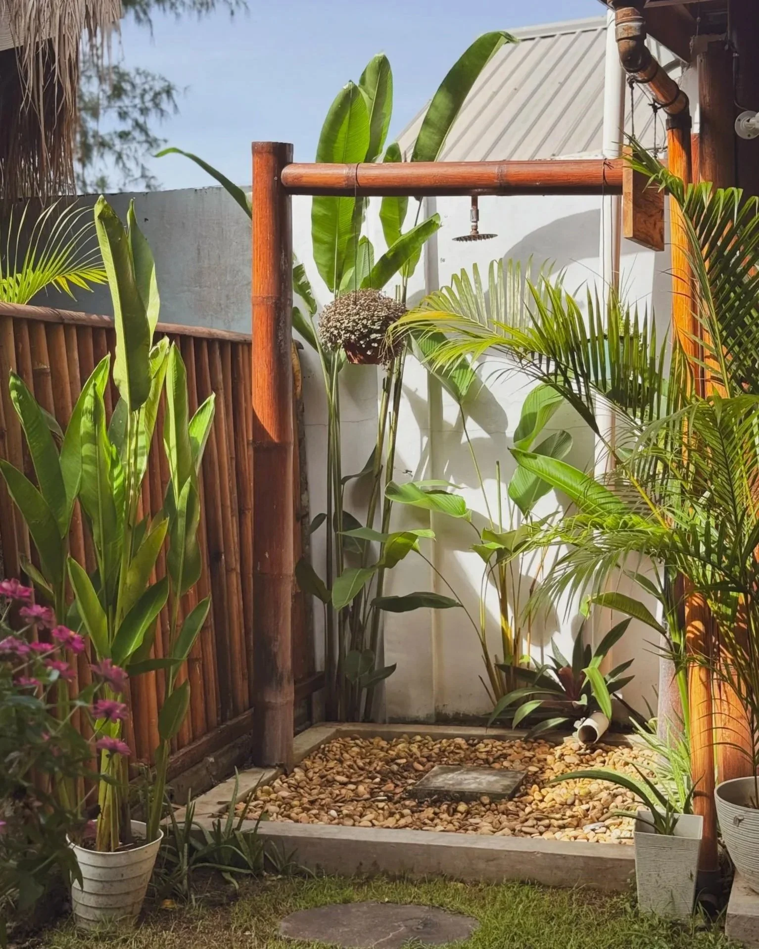 Outdoor shower area surrounded by lush green plants, with rocks on the ground and a white pipe on the right side.