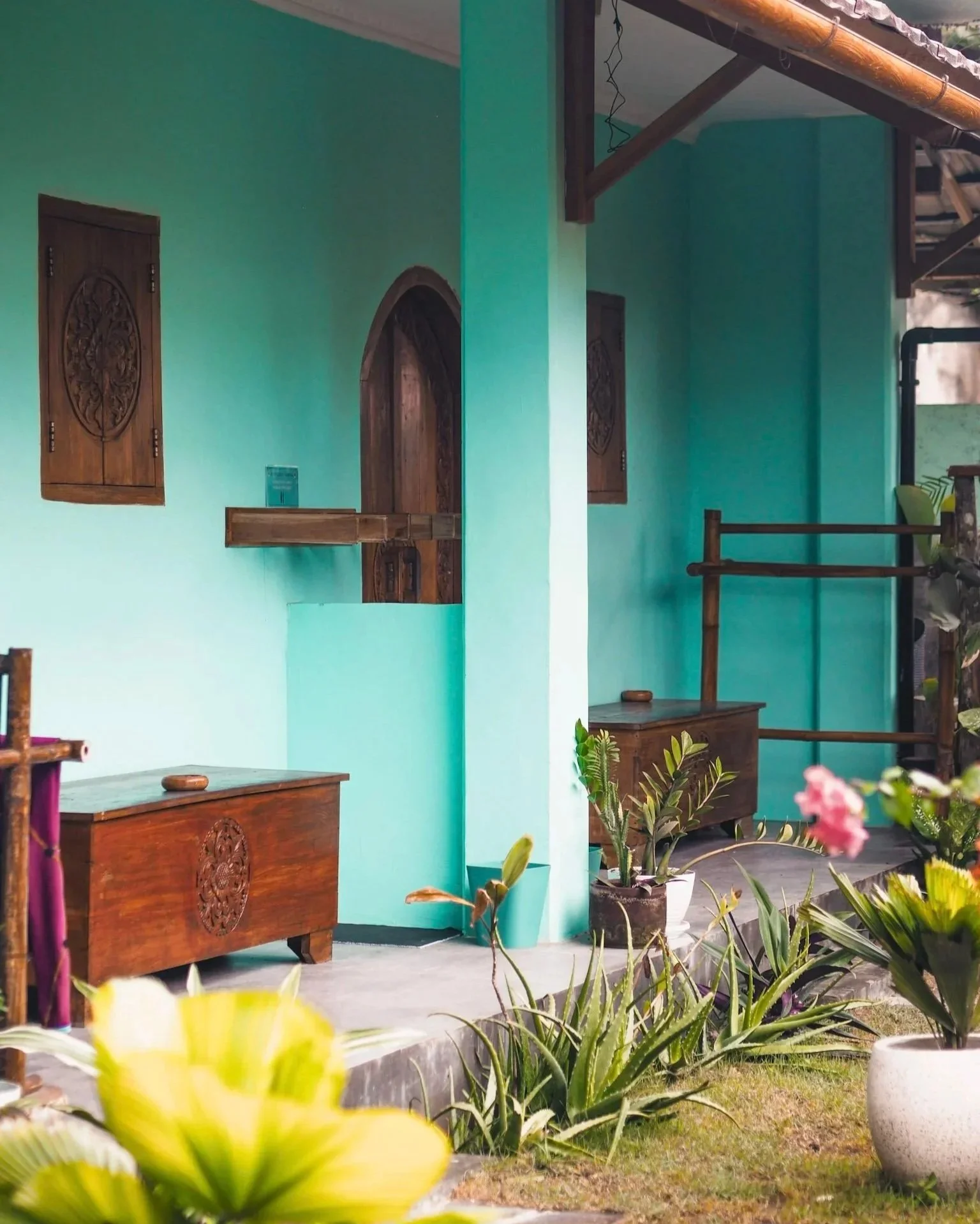 A brightly painted turquoise exterior of a small building with wooden decorative windows and furniture, potted plants, and an outdoor patio area.