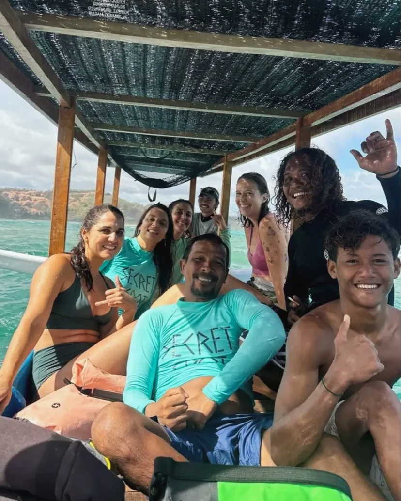 Group of eight people smiling and enjoying a boat ride on water with a scenic coastline in the background, under a shaded canopy.