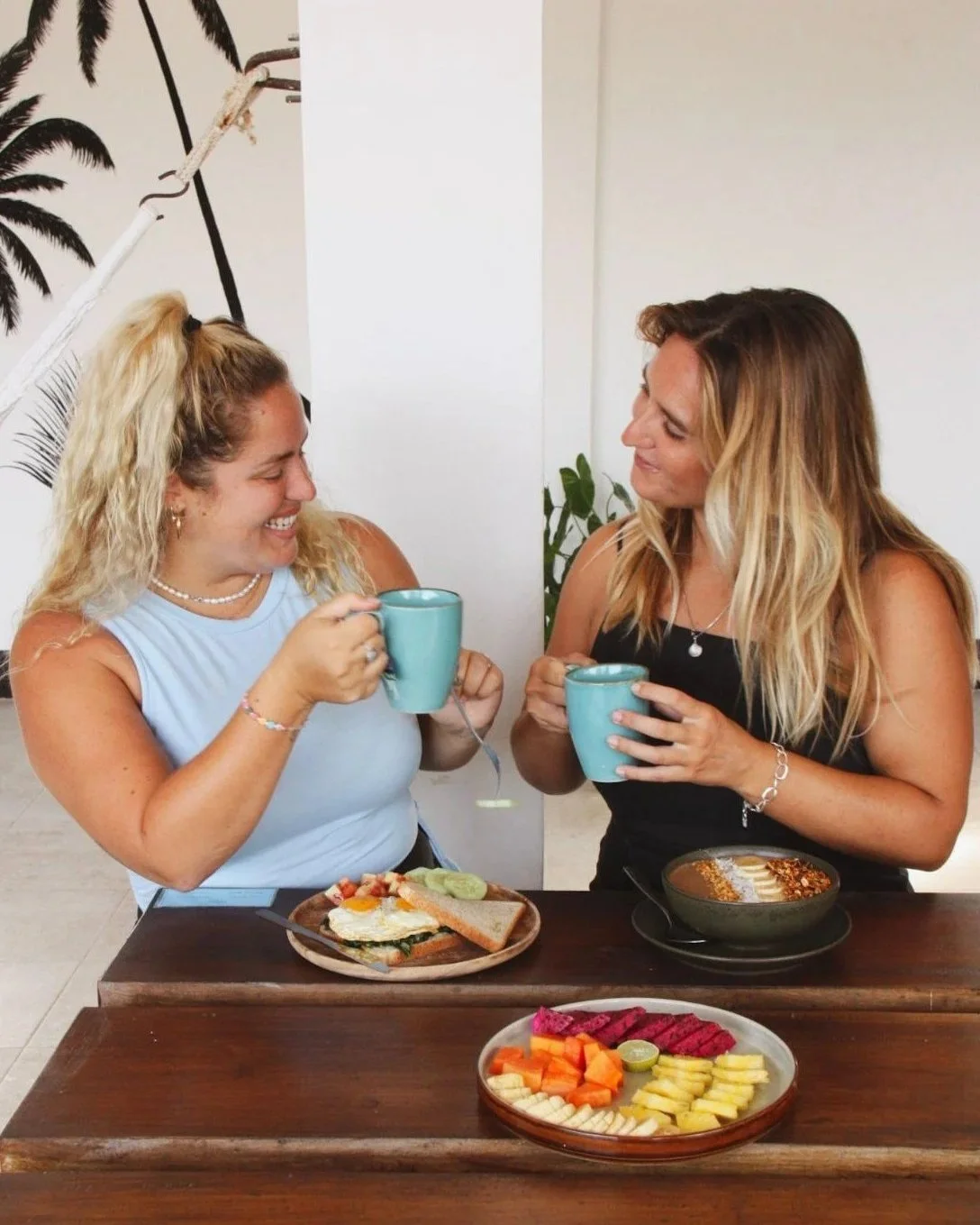 Two women smiling and holding blue mugs while having breakfast at a table with a plate of fruit and a bowl of cereal or granola.