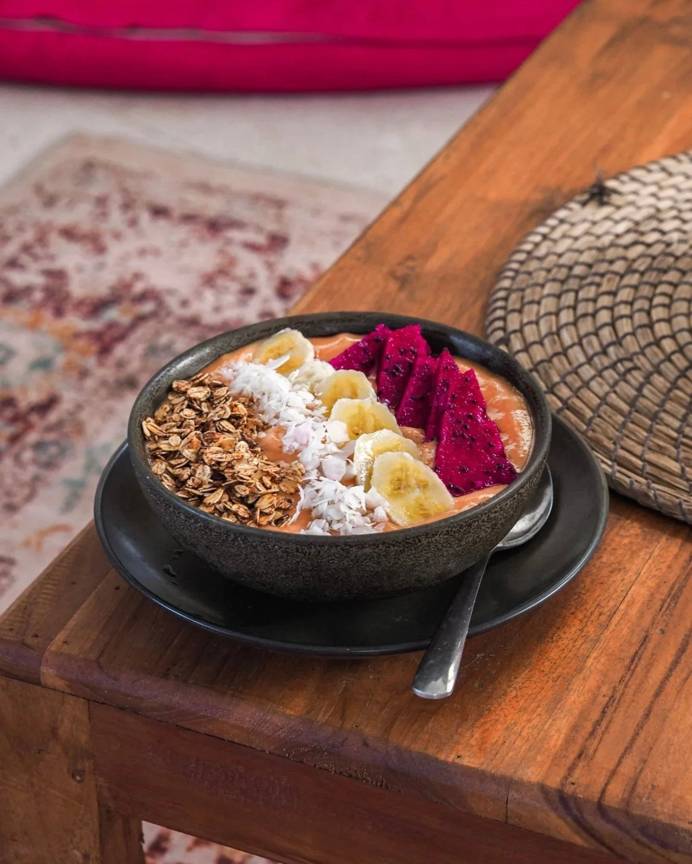 A bowl of smoothie topped with granola, sliced bananas, shredded coconut, and slices of dragon fruit, served on a black plate with a spoon.
