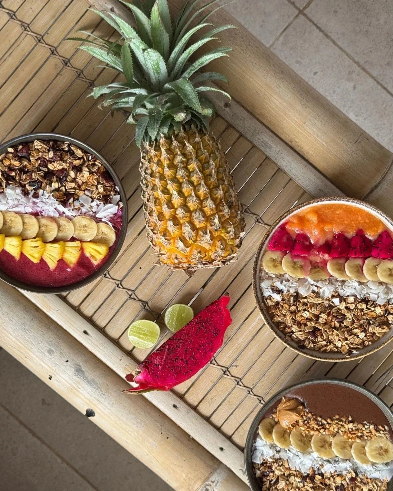 Three smoothie bowls with various fruits and toppings on a wooden table, with a pineapple and slices of lime and dragon fruit in the center.
