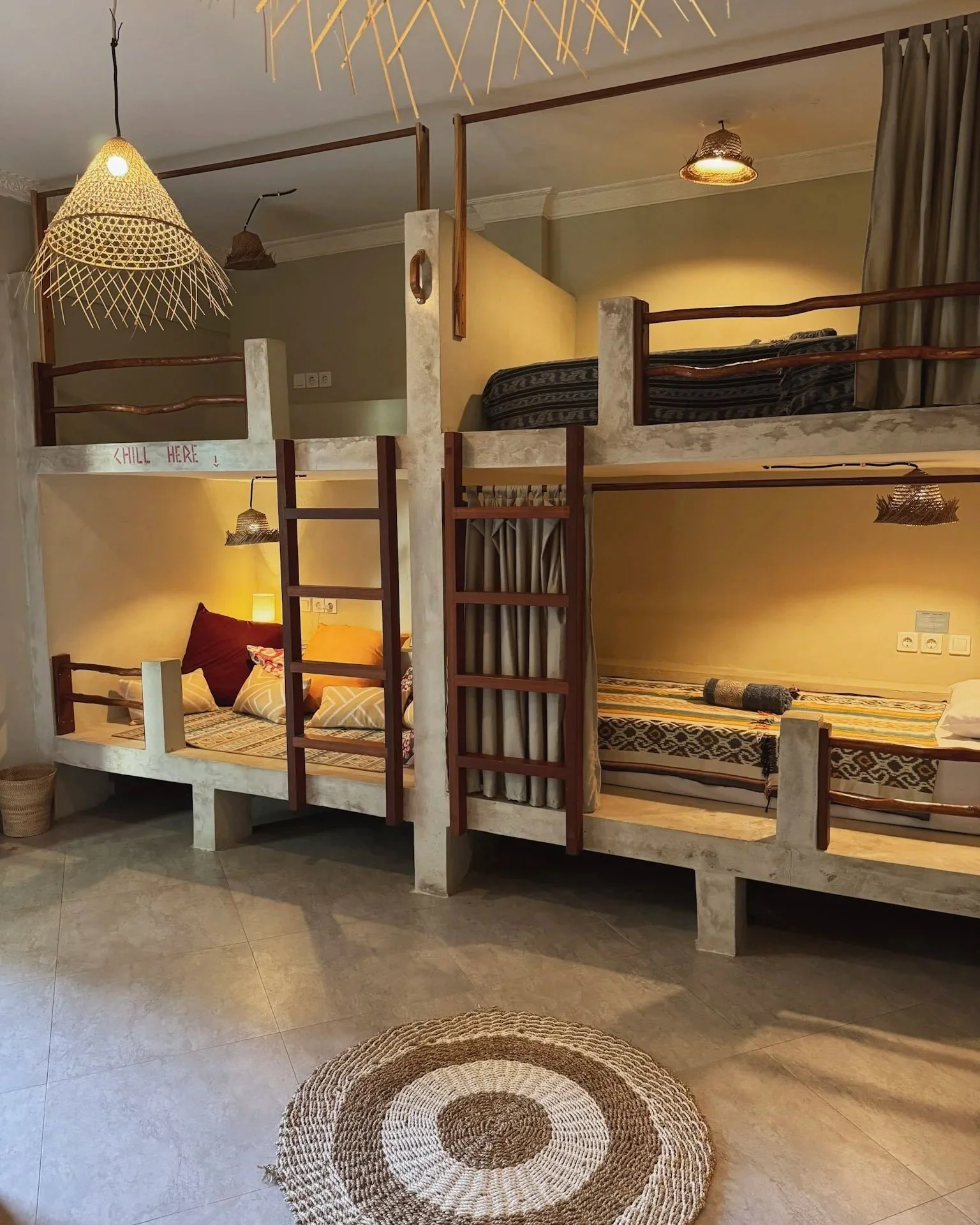 Interior of a hotel room with two bunk beds, each with a ladder, and warm lighting. The room has beige walls, patterned bedsheets, and woven lampshades hanging from the ceiling. A circular woven rug is on the floor.