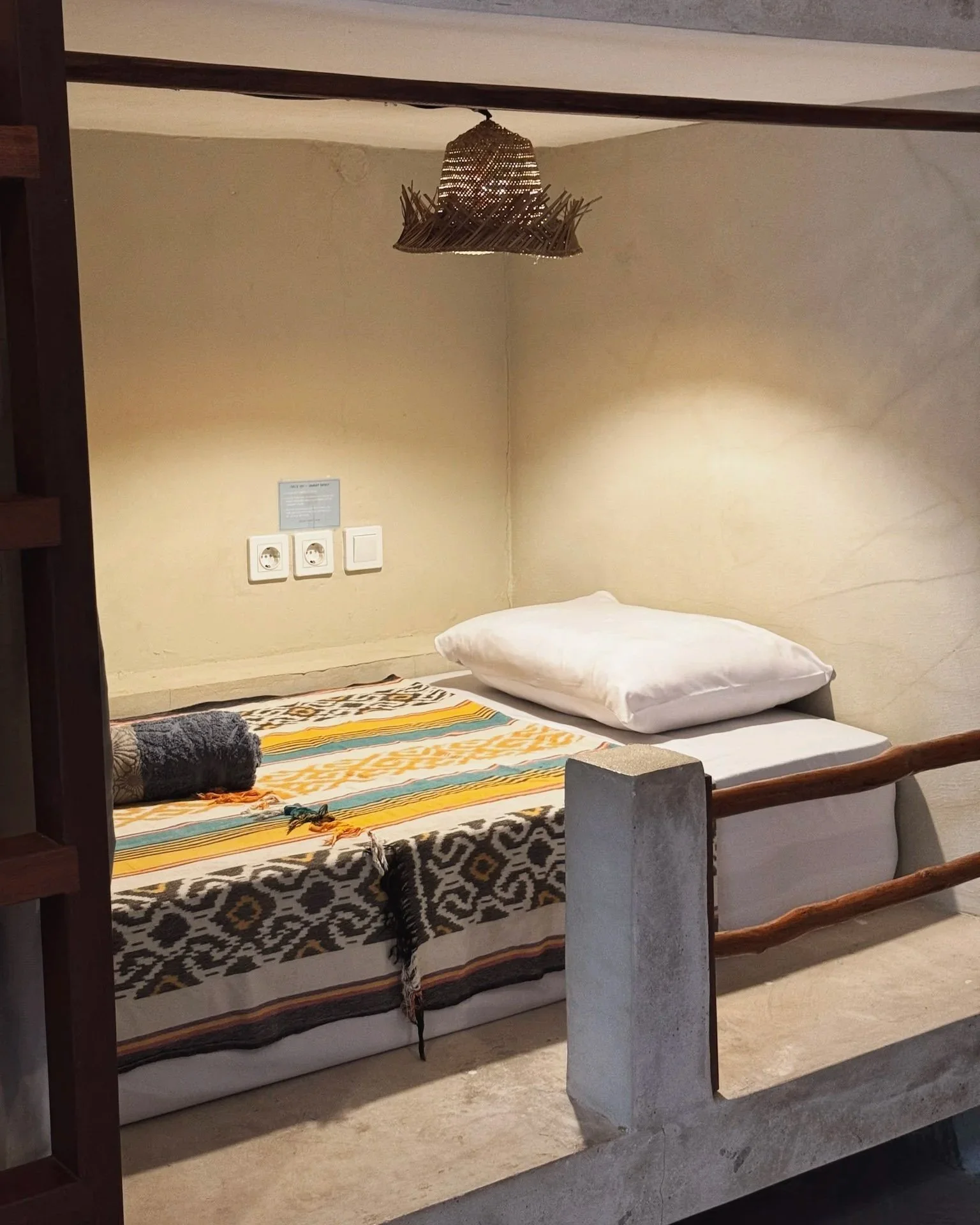 A bed with a white pillow and a colorful patterned blanket, situated on a small raised platform with a concrete railing. The wall behind the bed has electrical outlets and a blue information sign, with a woven wicker lampshade hanging from the ceilin
