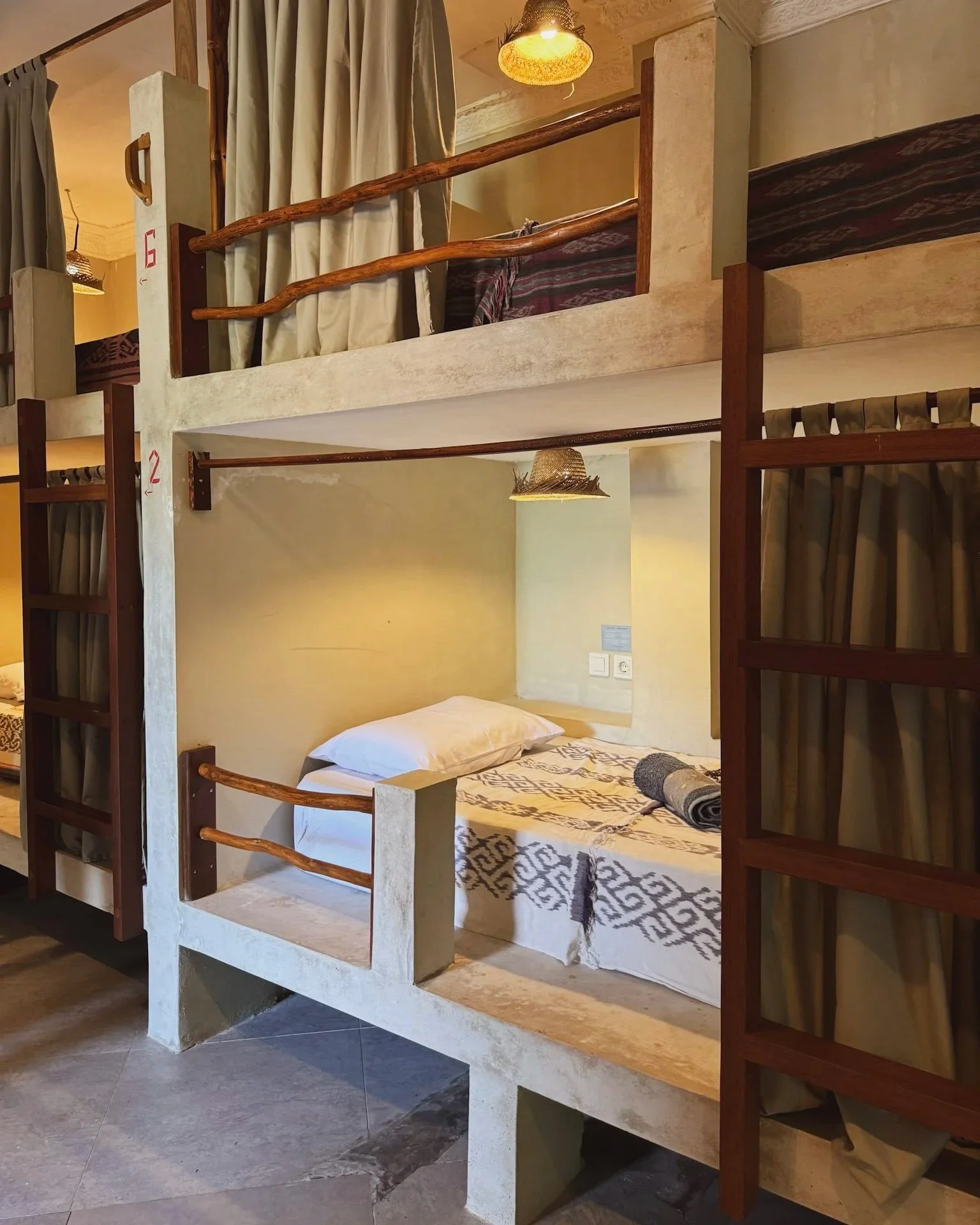 Bunk beds with curtains in a cozy, rustic style hotel room, featuring a lower bed with a rolled towel and a pillow, and an upper bed with privacy curtains, wooden railing, and decorative lighting.