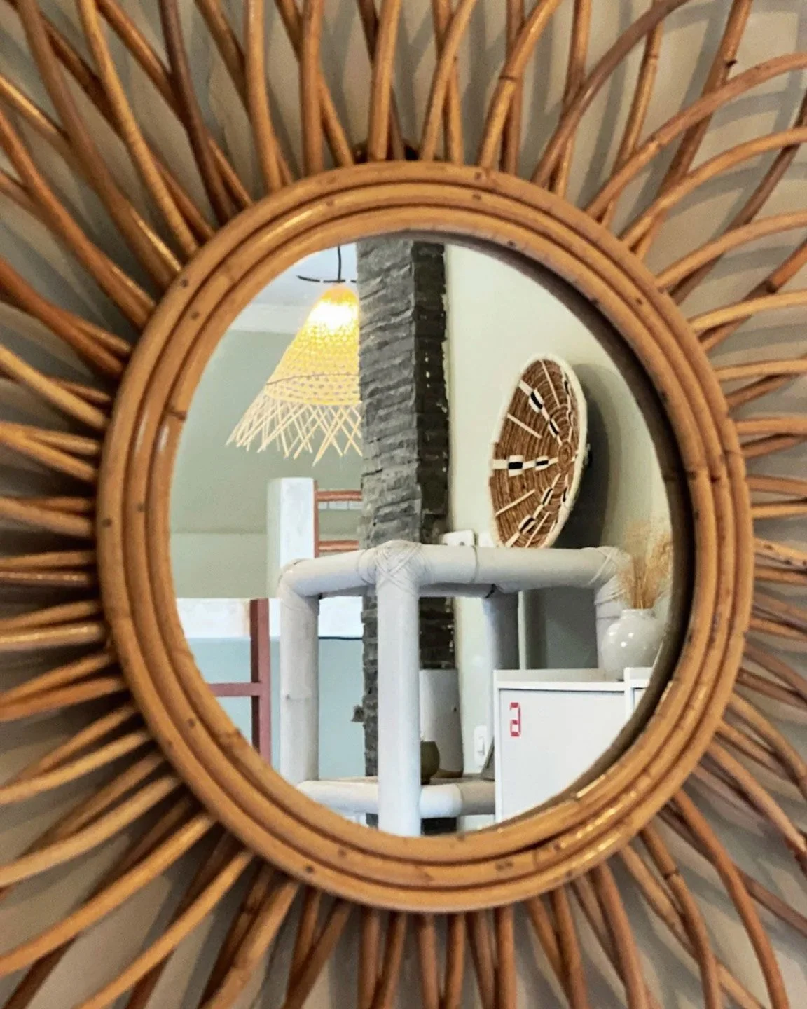 A round mirror with a wicker frame, reflecting part of a room with a lamp, a wall clock, and some furniture.