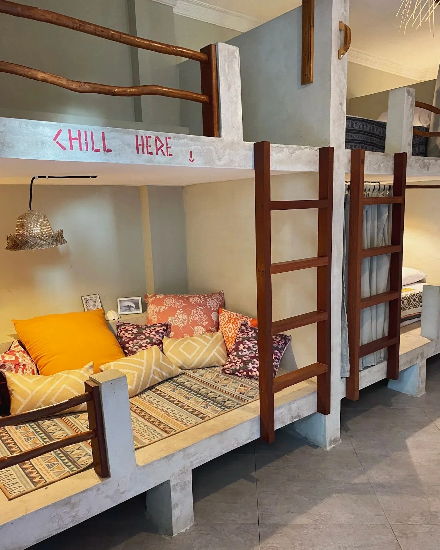 Interior of a colorful room with built-in seating and a loft area. The bottom area has cushions and pillows, some with floral and geometric designs, and a patterned mat on the seat. The loft is accessible by small wooden ladders and has a sign that r