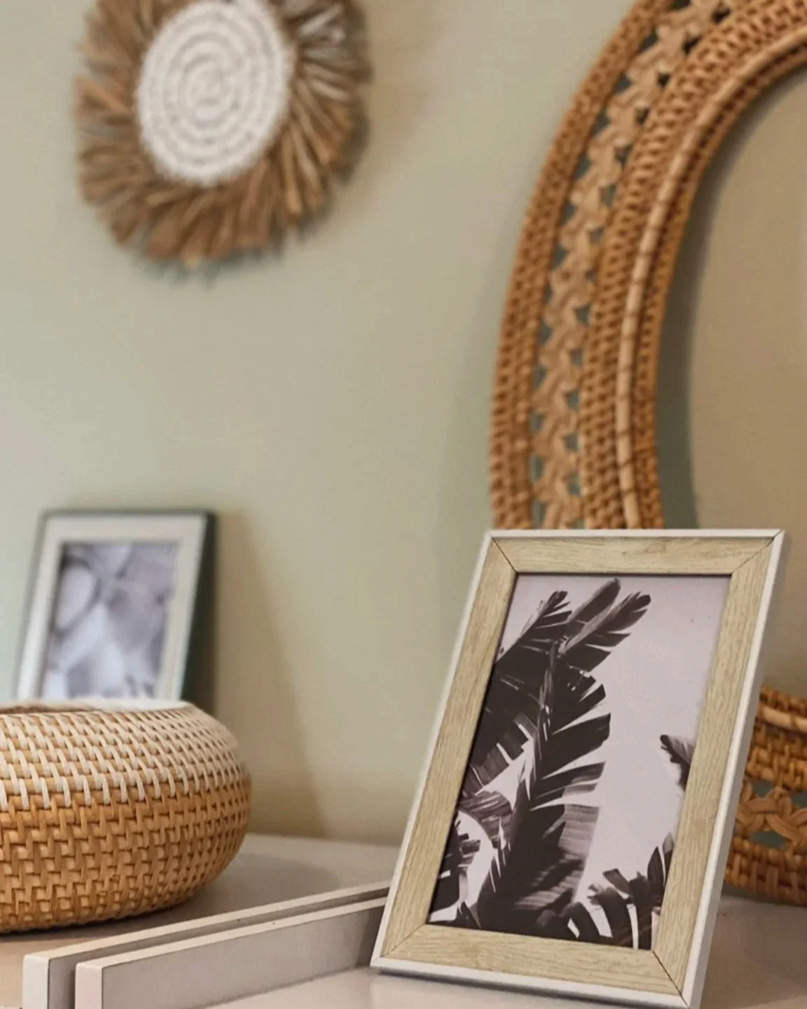 A black and white photo of tropical leaves in a wooden frame, part of a home decor arrangement on a white table, with woven baskets and wall decorations in the background.
