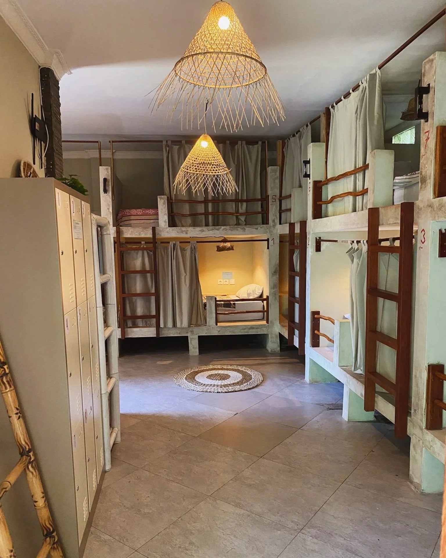Interior of a hostel with bunk beds and individual privacy curtains, wooden ladders, two woven pendant lights, beige lockers, and a round rug on tiled floor.