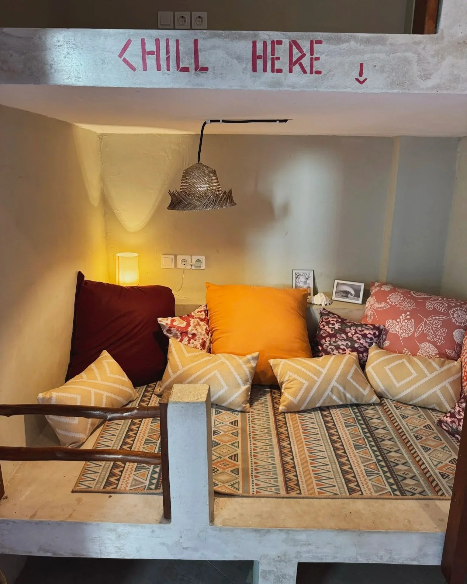 Cozy seating area with pillows on a patterned rug, a small lamp, framed artwork, and a hanging lamp in a room with textured walls and a concrete ledge with 'CHILL HERE' painted on it.