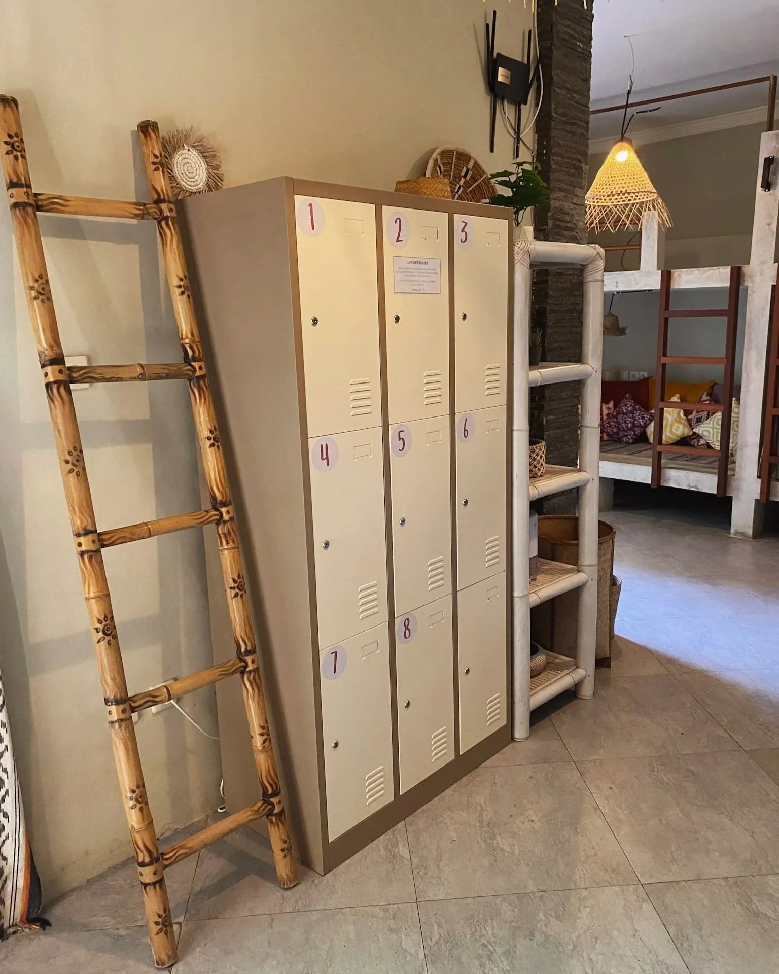 A row of small beige lockers with numbered pink and white circular tags from 1 to 8, placed against a beige wall. To the left of the lockers is a bamboo ladder with floral designs on the rungs, leaning against the wall. To the right of the lockers, t