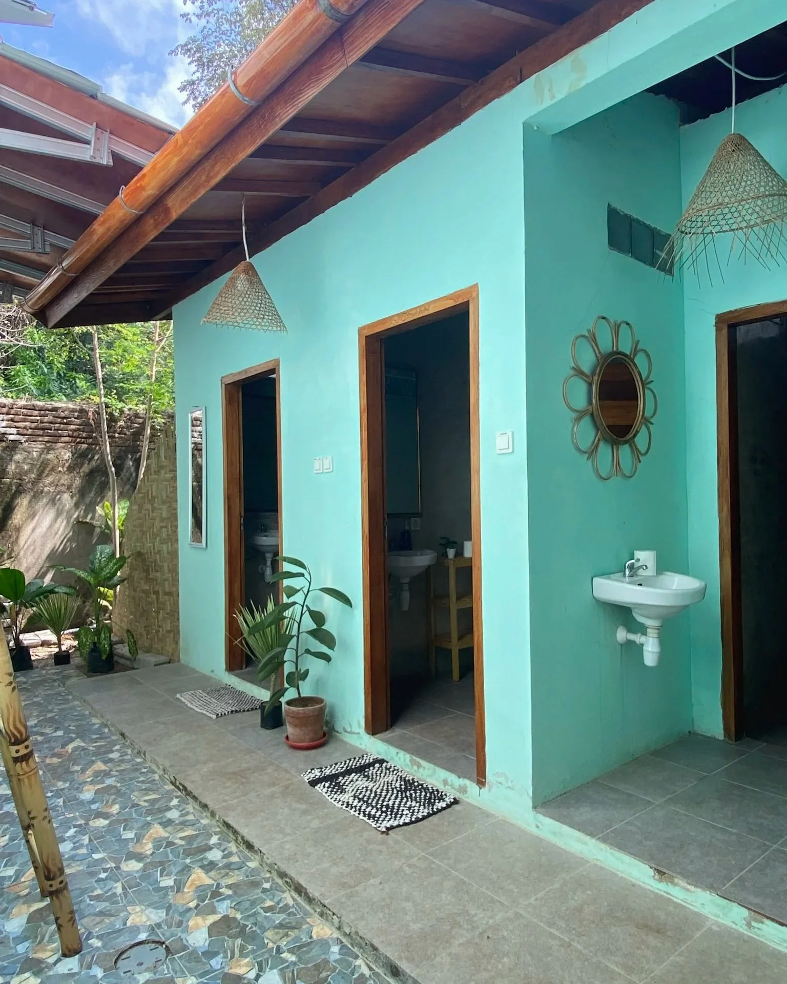 The image shows a turquoise-colored building with three open doorways, each leading to indoor spaces. There are two small bathrooms visible, with a sink outside one of them. The exterior area has a patterned stone pathway, potted plants, and two wove