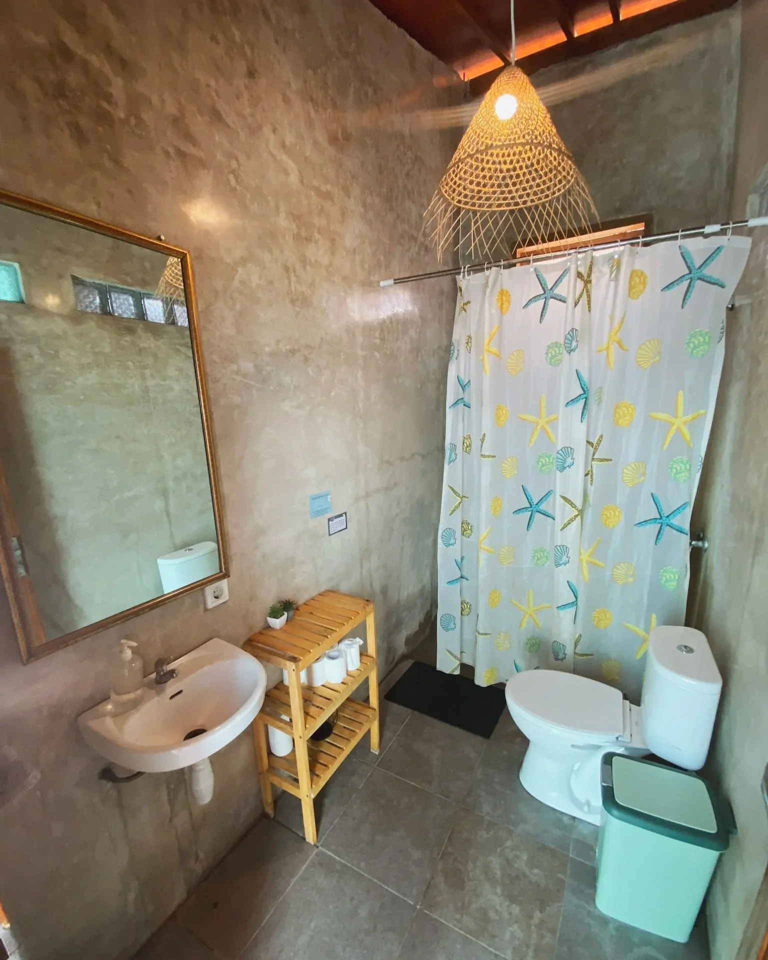 Bathroom with concrete walls, a mirror, a small sink with soap, a wooden shelf with toilet paper rolls, a toilet with a colorful starfish and shell shower curtain, a small trash bin, and a woven hanging light fixture.