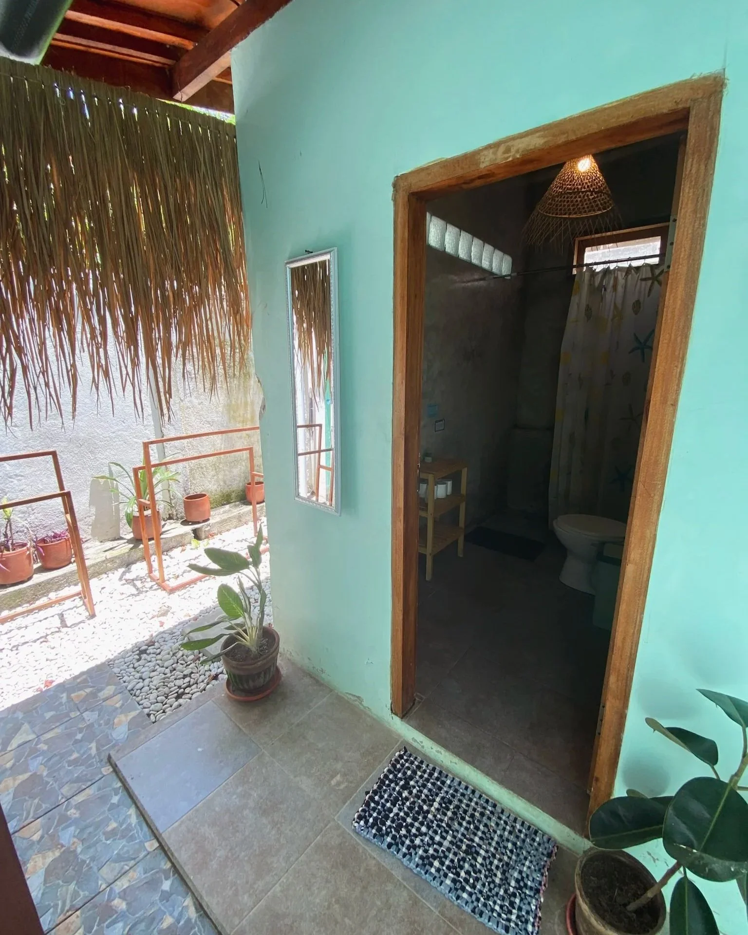 View from a porch area with a door leading to a bathroom. The porch has tiled flooring, potted plants, and a thatched roof overhang. The bathroom interior is dimly lit, with a shower curtain and a toilet visible.