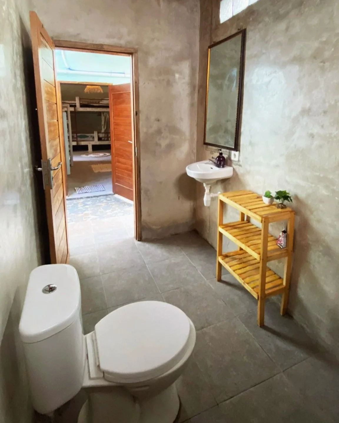 A rustic bathroom with concrete walls, a toilet, a small white sink with a mirror above it, a wooden shelf with plants and toiletries, and a door leading to an adjacent room. There is a skylight with glass blocks and a large woven decor piece hanging