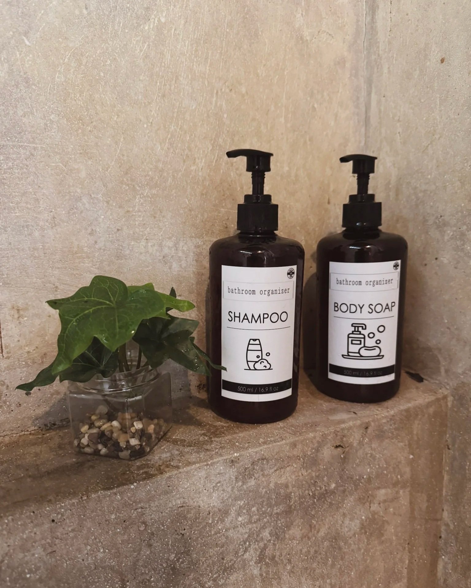 Two dark bottles with pump dispensers labeled 'Shampoo' and 'Body Soap' sitting on a stone shelf against a beige tiled wall. A small potted ivy plant in a clear container with pebbles.