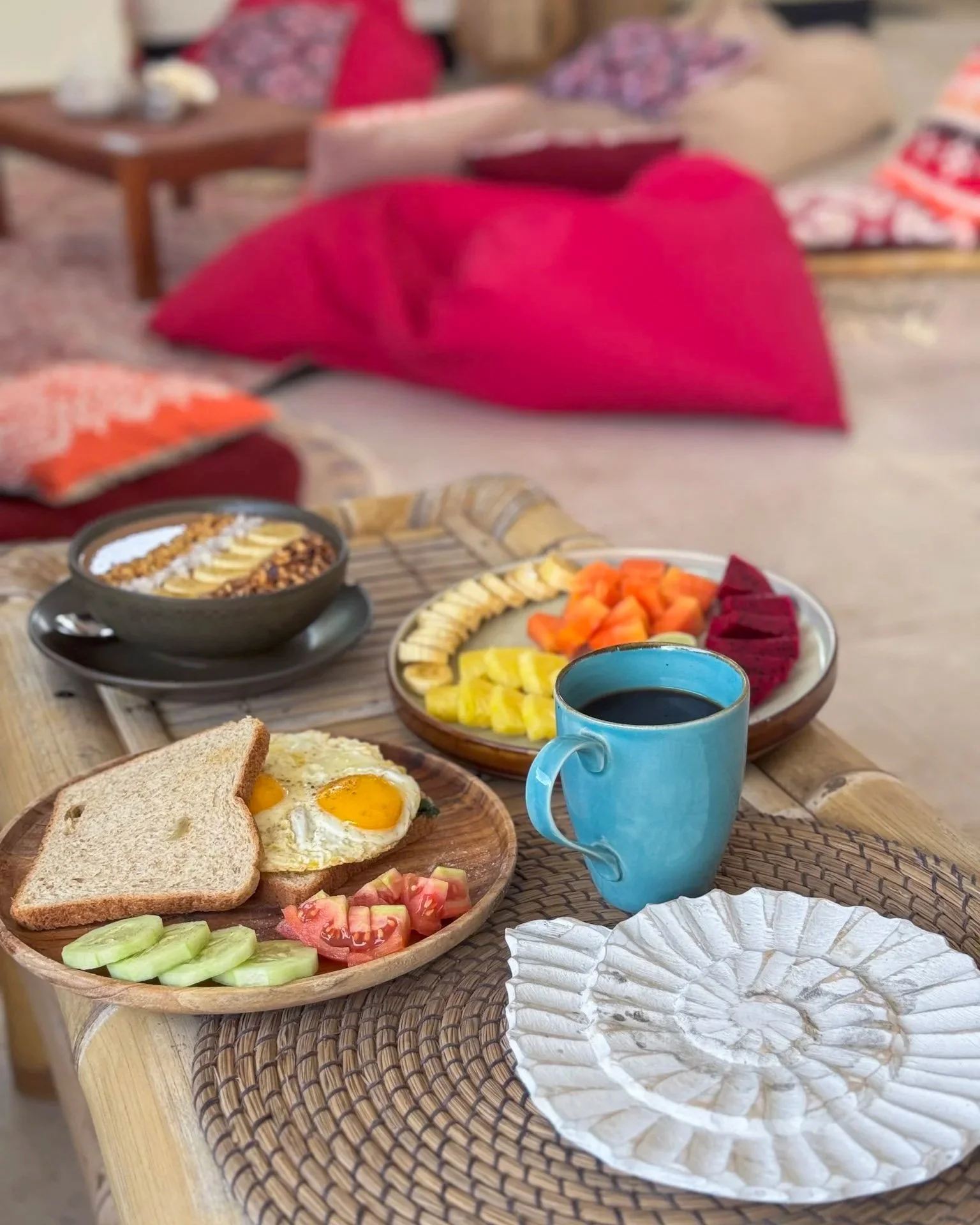A breakfast setup on a wooden table with a plate of sunny side up eggs, toast, sliced cucumbers, and tomatoes; a bowl of cereal with dried bananas; a plate of sliced fruits including bananas, papaya, pineapple, and dragon fruit; a cup of black coffee