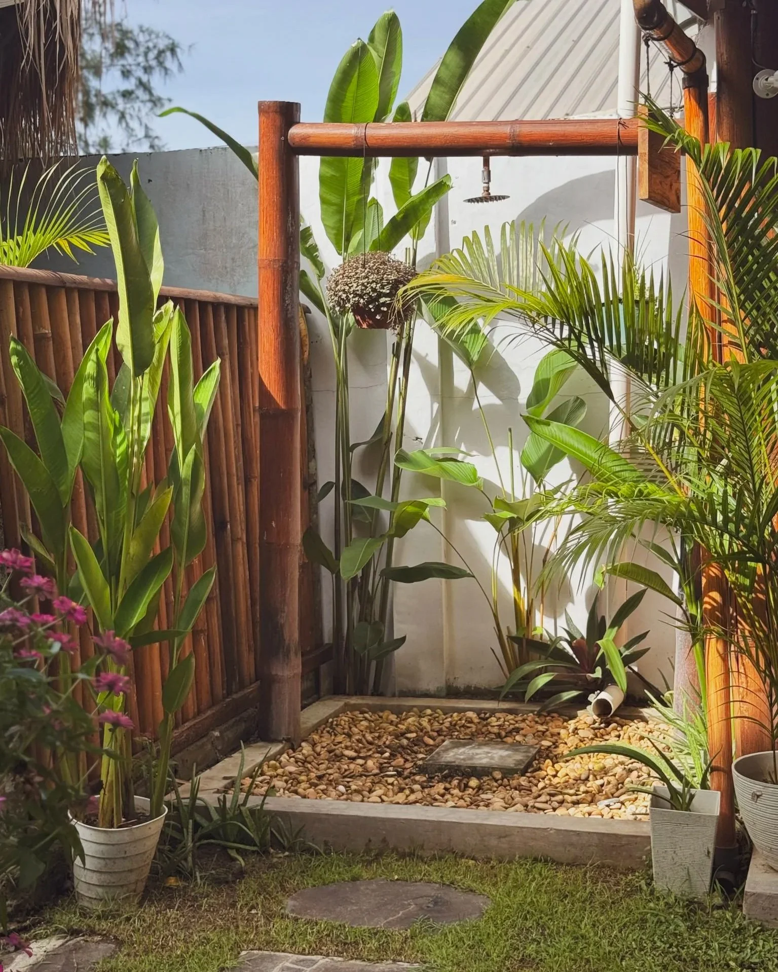 A small outdoor shower area with a wooden frame and a rain shower head, surrounded by lush tropical plants and potted flowers, with a pebble-filled base and a small stepping stone.