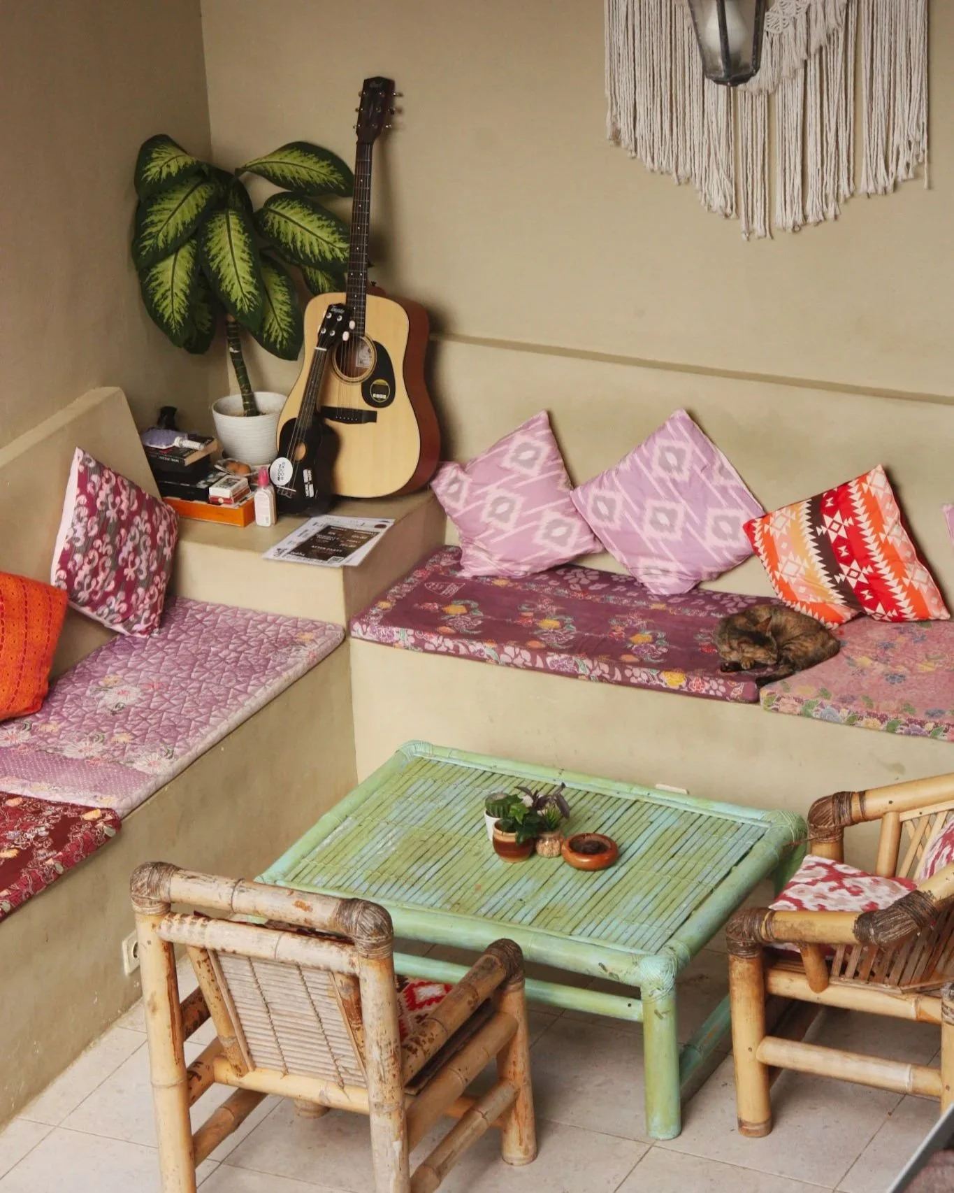 A cozy living room corner with a built-in couch decorated with pink, red, and floral cushions, a green coffee table with potted plants, two rattan chairs, a guitar resting against a large houseplant, a small table with some items and a cat curled up 