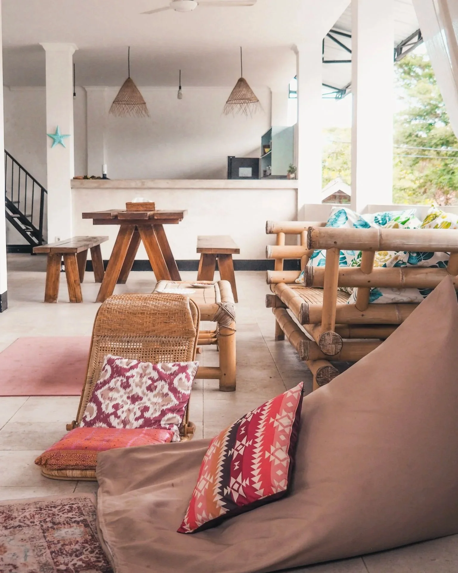 A bright, airy living room with tropical-inspired furniture, including a bamboo sofa with colorful cushions, a woven chair with a pink patterned pillow, and a wooden bench. There's a dining area with a wooden table, benches, and hanging woven light f