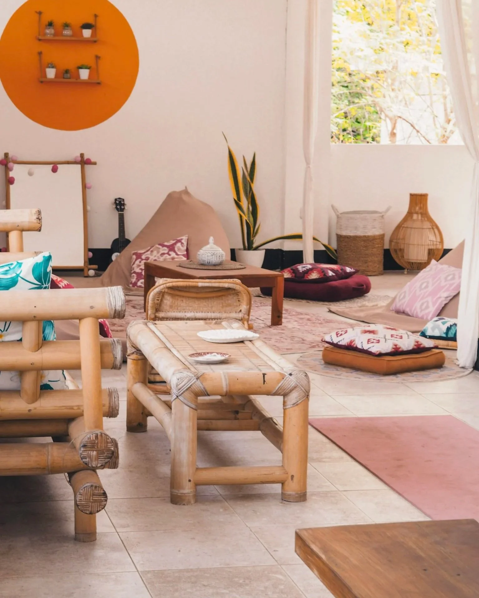 A cozy indoor space with bohemian decor, including bamboo furniture, pillows, rugs, and houseplants, near a window with white curtains letting in natural light.