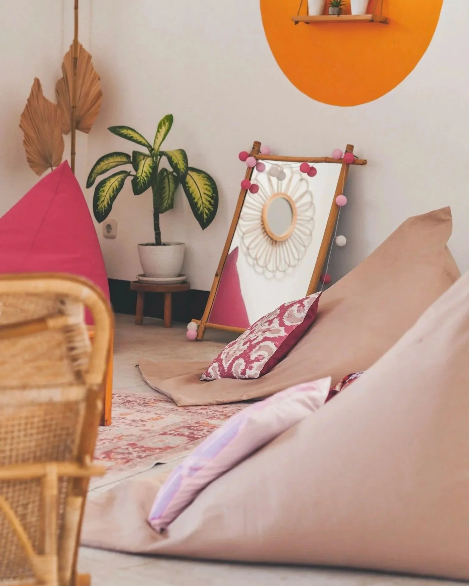 Bohemian-style cozy living room with large pink and beige floor cushions, a rattan chair, a potted plant, a mirror with a floral frame, and a wall shelf with small potted succulents.